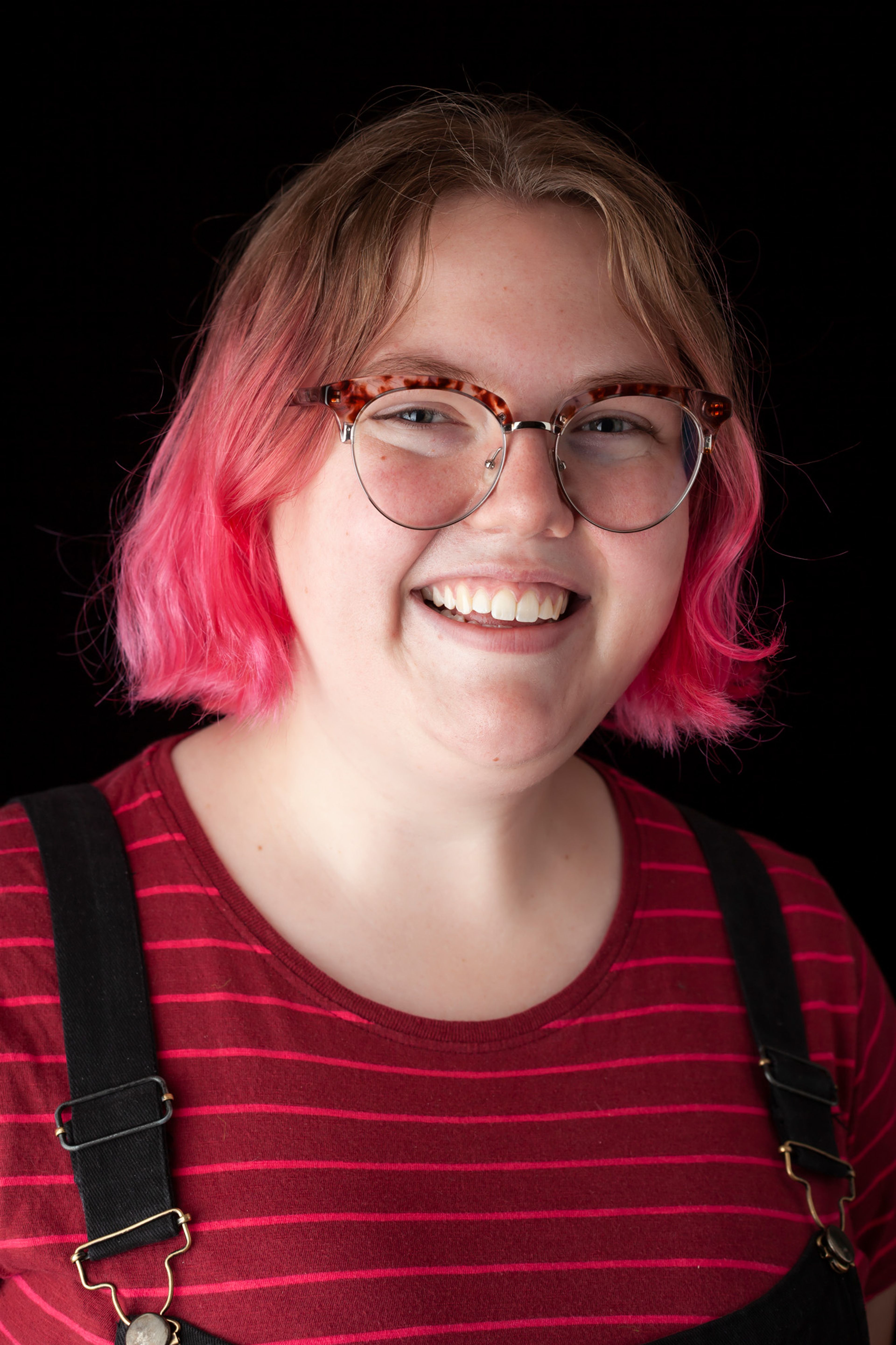 A portrait of a smiling individual with short pink hair, wearing tortoiseshell glasses, a red striped shirt, and black overalls