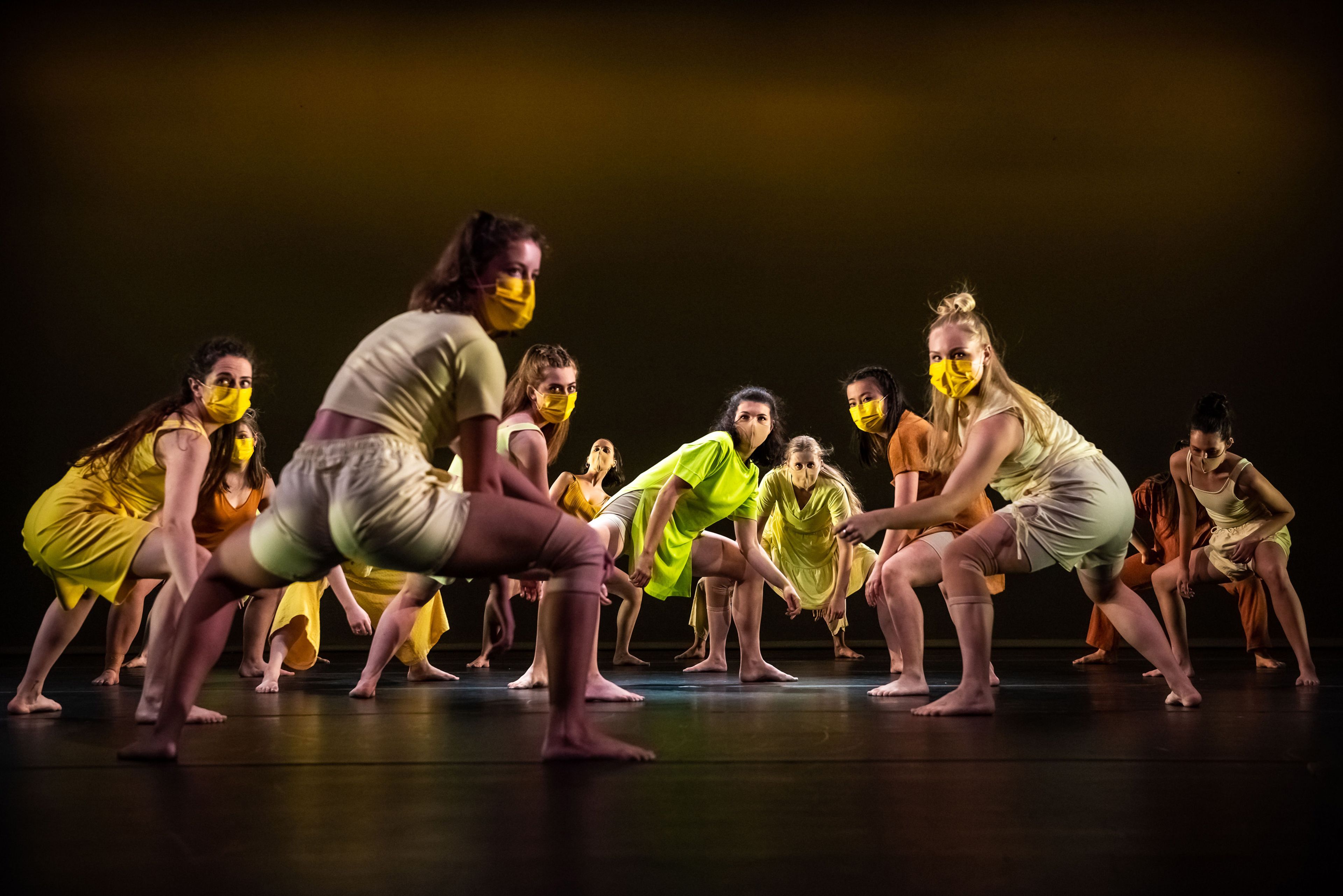 A group of dancers crouched in low stances on stage wearing yellow and orange outfits snd matching face masks. The scene is lit warmly