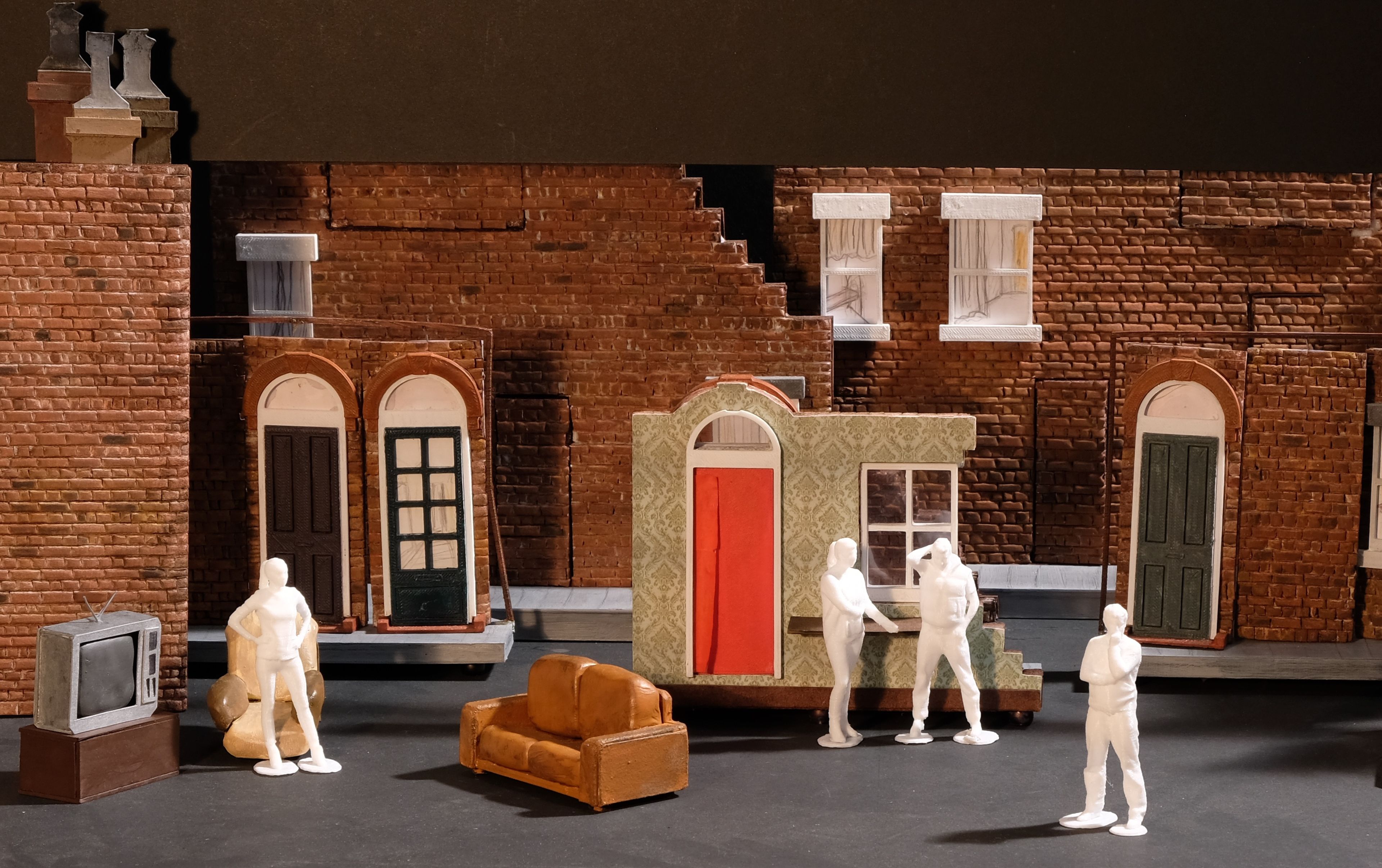 A model box of stage design with red-brick facades, doors, windows, and white figurines posed interactively. Props include a vintage TV, sofa, armchair, and a freestanding wall with a red door