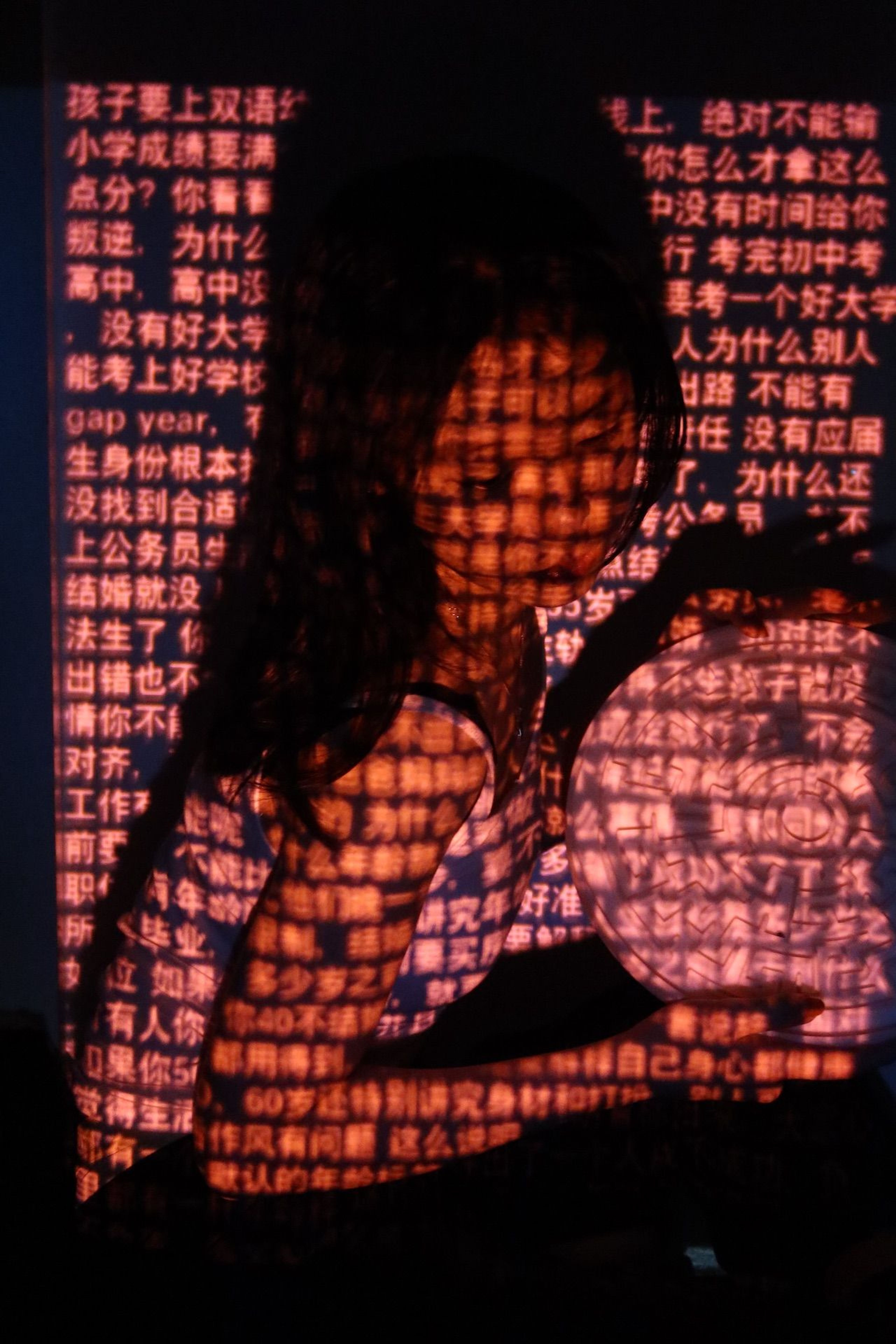 Photo of a dark-haired girl in a white singlet and black pants crouching in the dark, holding a wheel with a labyrinth print, with projection of rows of red Chinese characters.