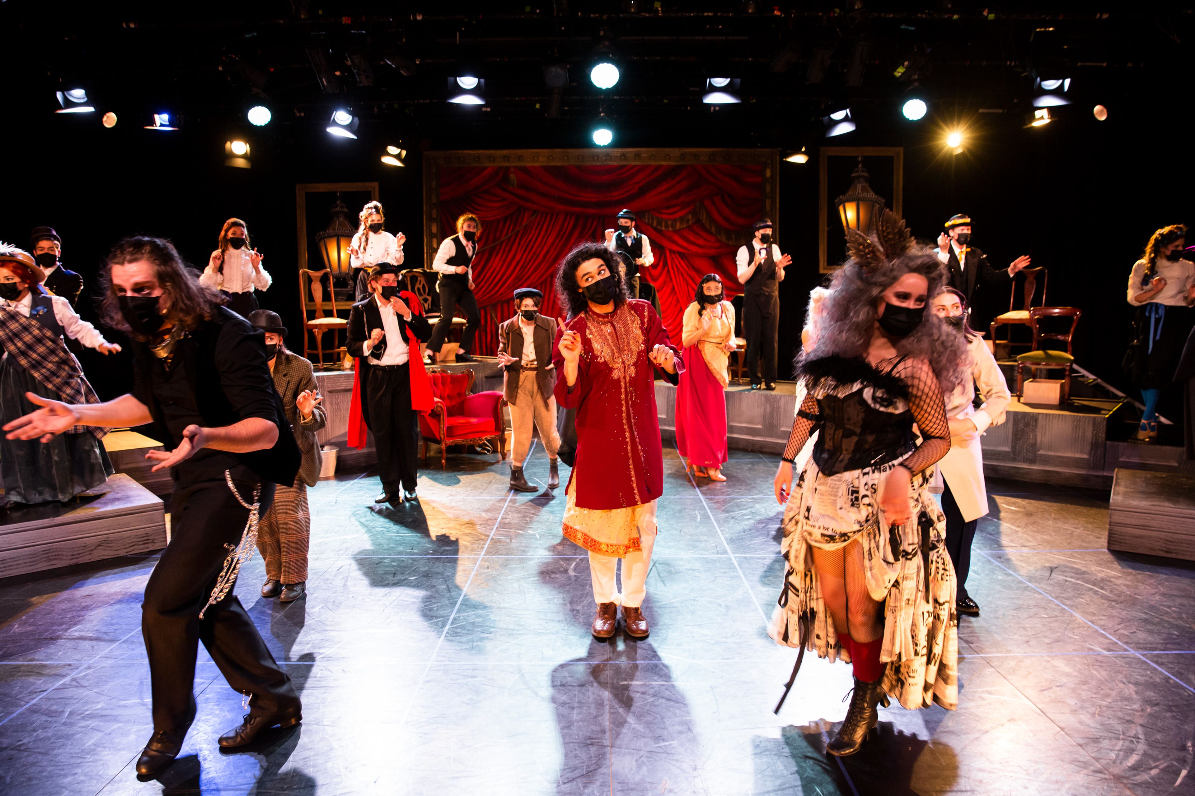 A theatrical performance featuring a large ensemble of performers dressed in elaborate costumes and face masks, standing and moving across a brightly lit stage with a red curtain backdrop