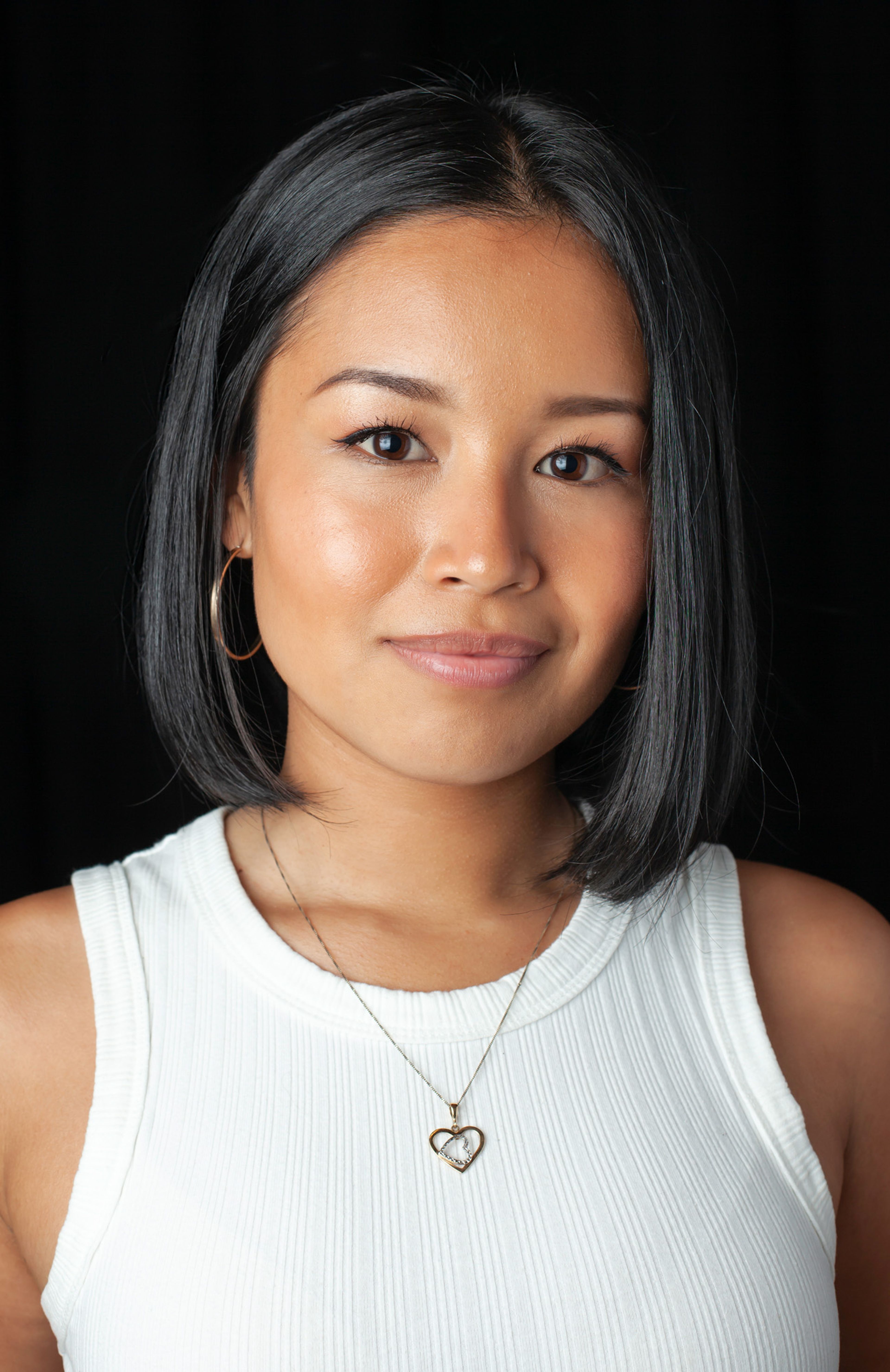 A woman with a sleek bob, white top, heart necklace, and gold hoop earrings, set against a black background