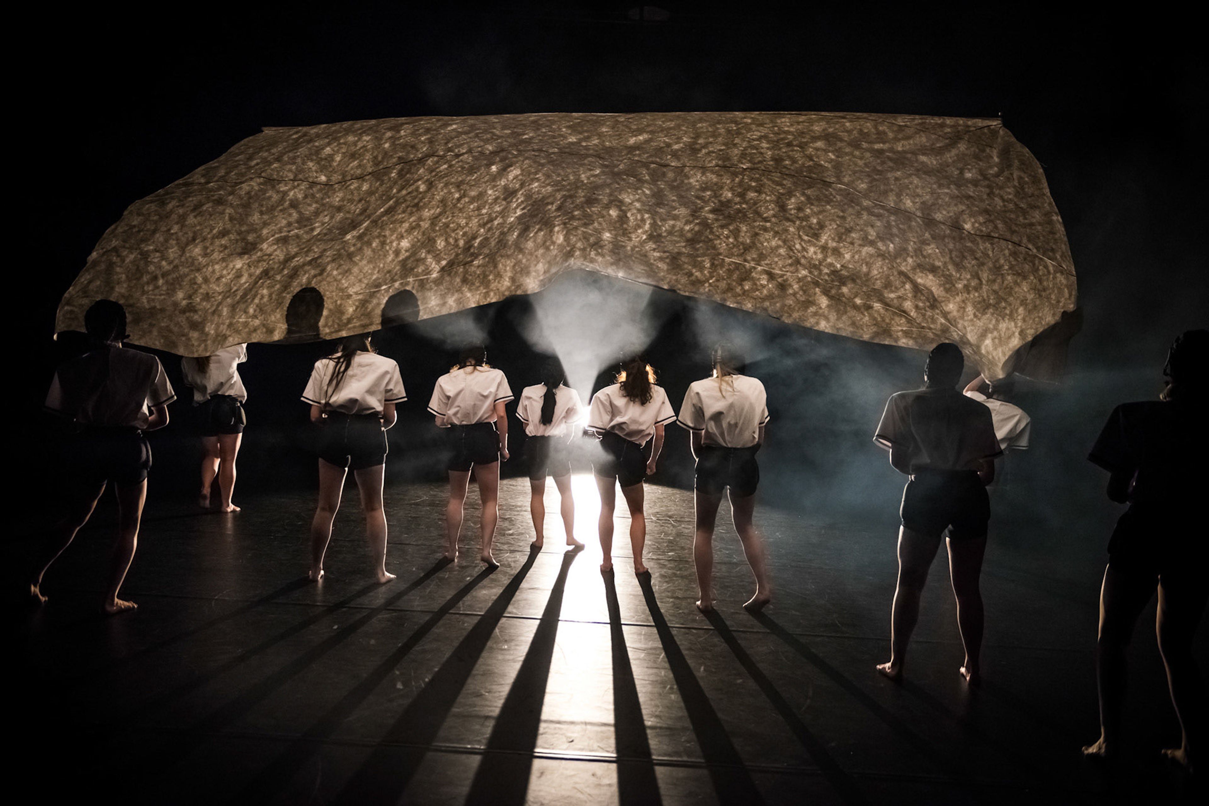 A group of performers stands in a dimly lit space, silhouetted against a glowing light source behind them, creating dramatic shadows