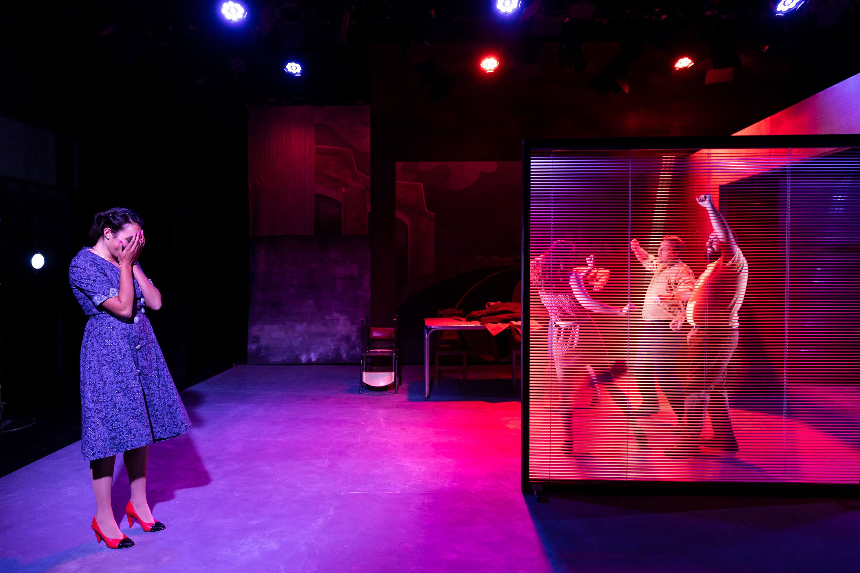 A woman in a blue dress and red shoes covers her face in distress, while three others dance energetically behind blinds under red and purple lighting