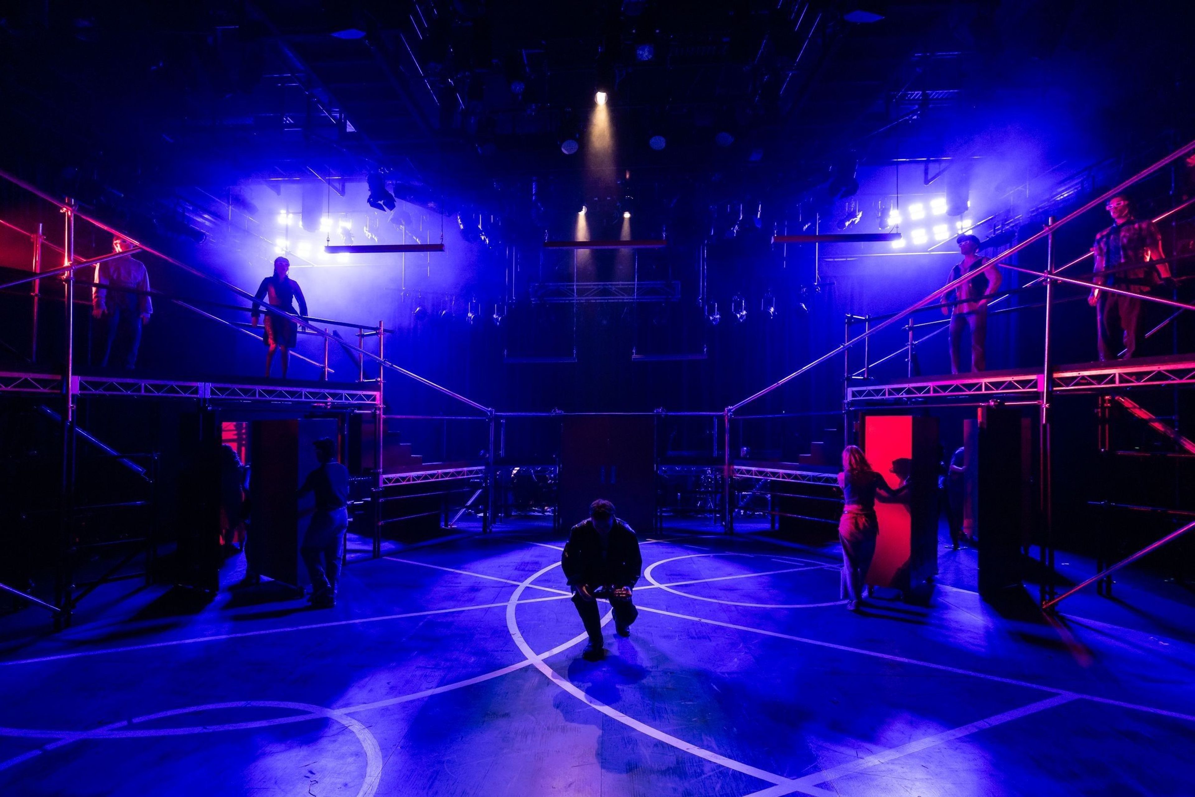 Theatre set with scaffolding and actors under blue and red stage lighting.