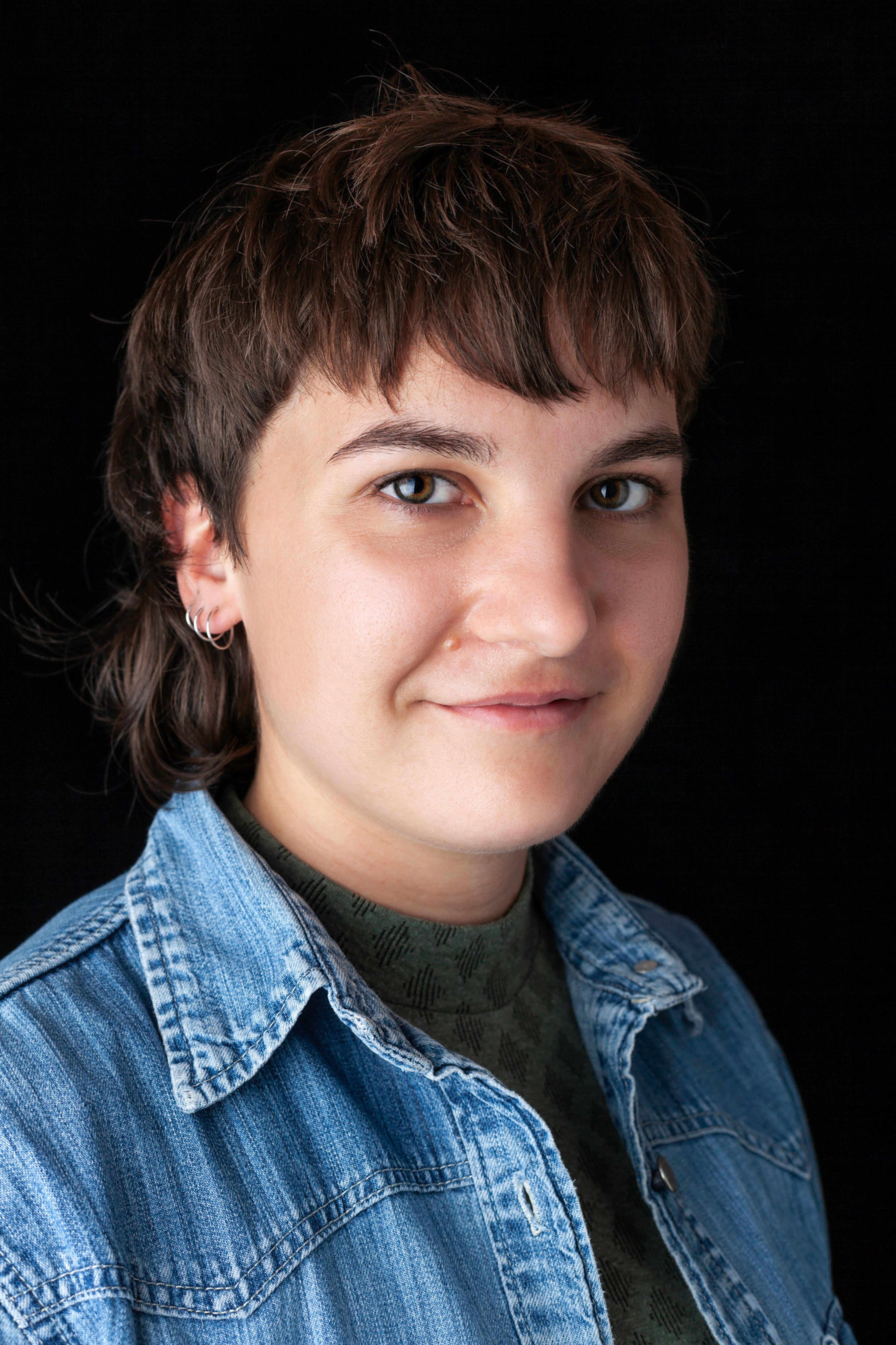 A portrait of a person with short, textured brown hair, wearing a denim jacket. They have a slight smile and are wearing small silver hoop earrings