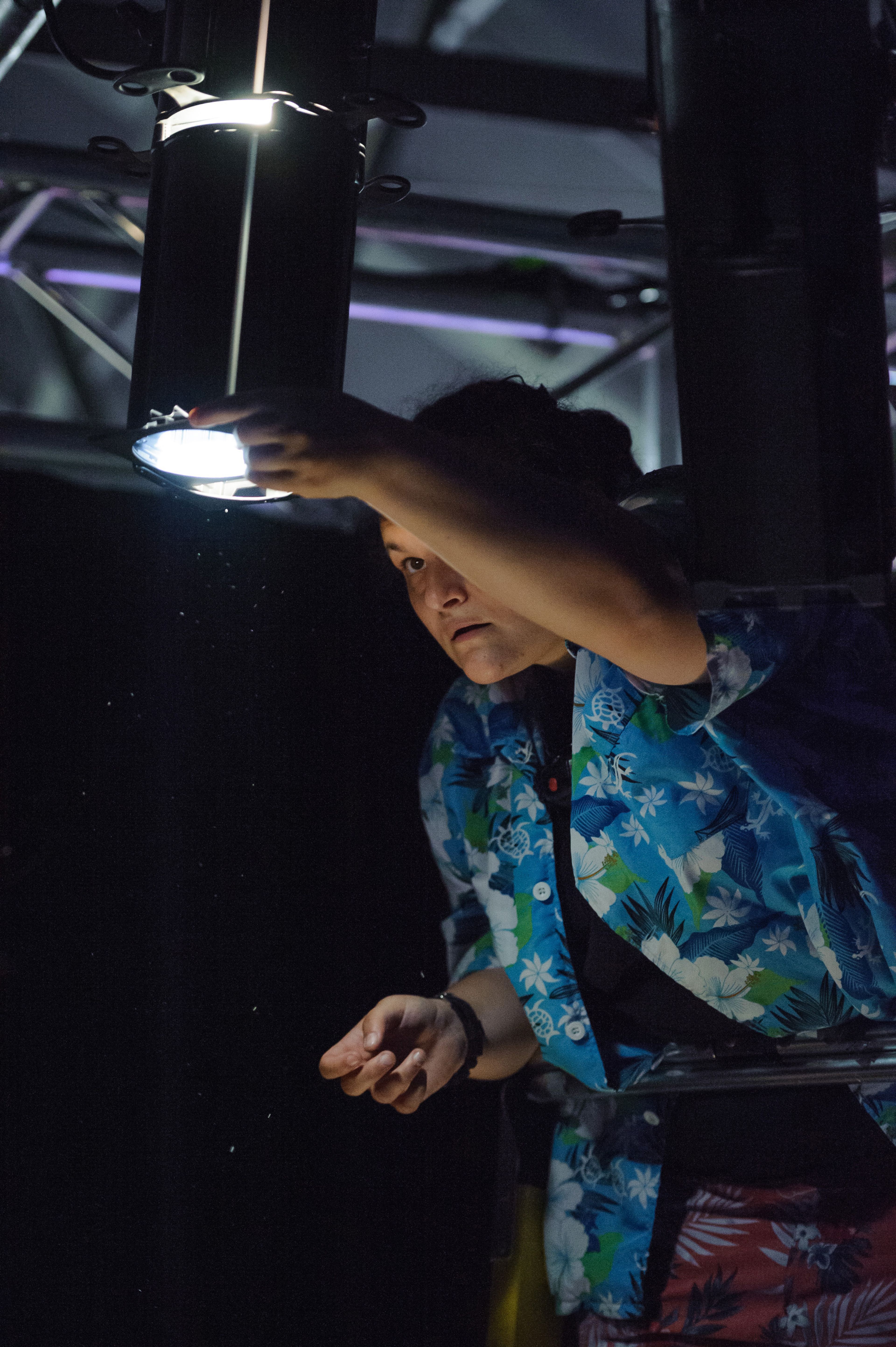 A person adjusts a stage light backstage, focused and precise in a dimly lit setting