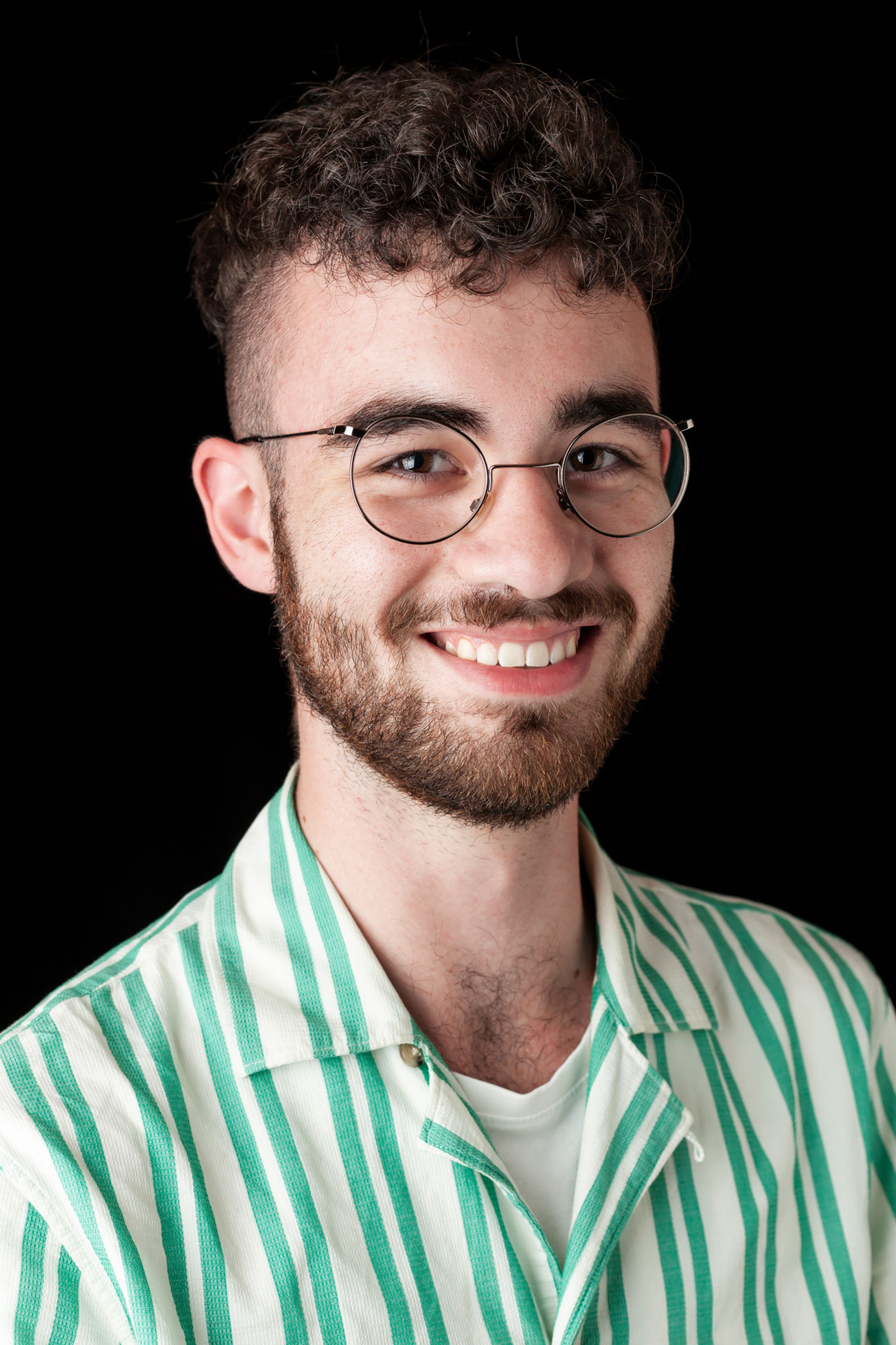 A portrait of a smiling man with curly hair, glasses, and a beard, wearing a green and white striped shirt