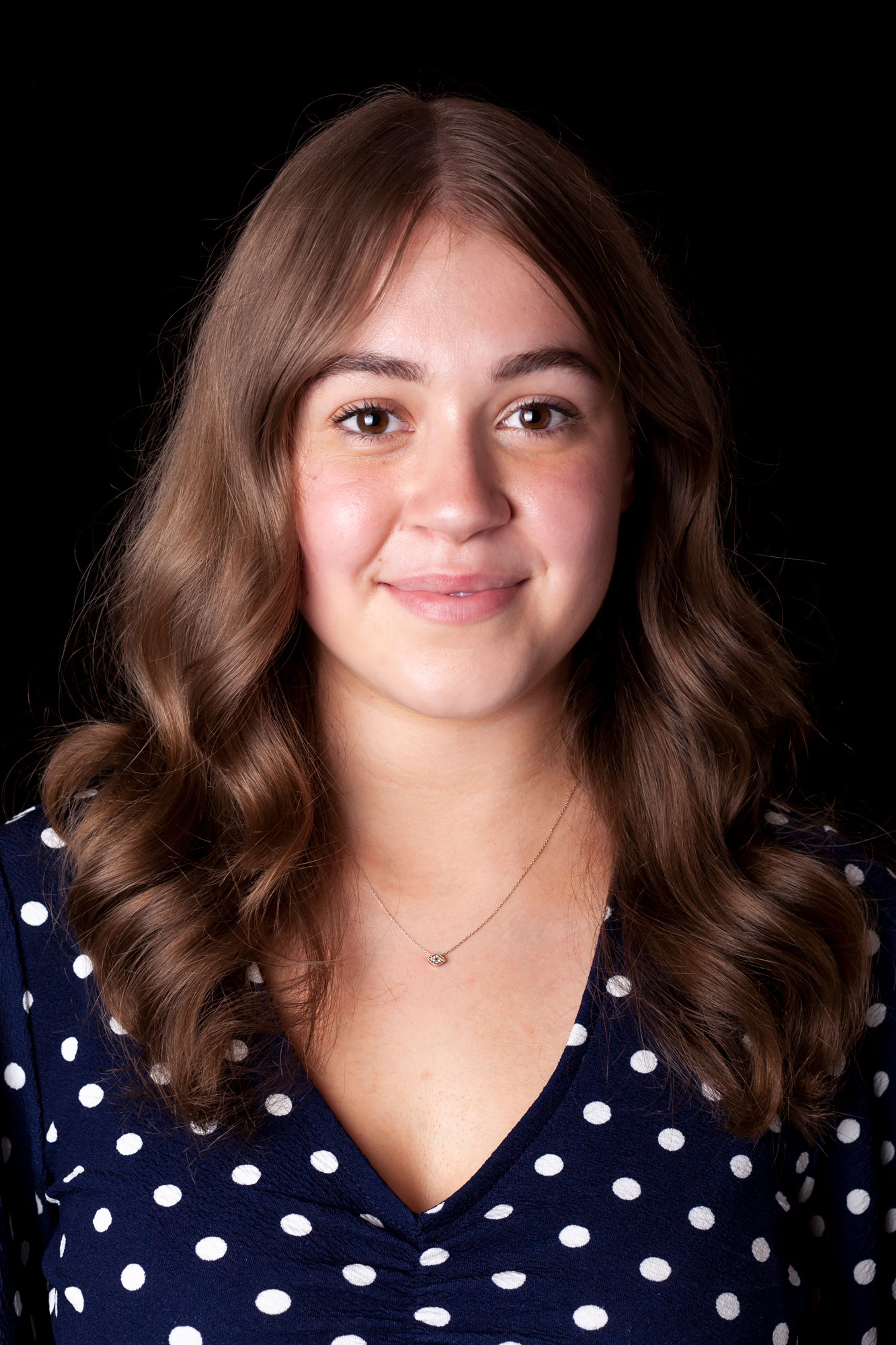 Photograph of a smiling woman with wavy brown hair, wearing a navy blue polka-dot top and a delicate necklace 