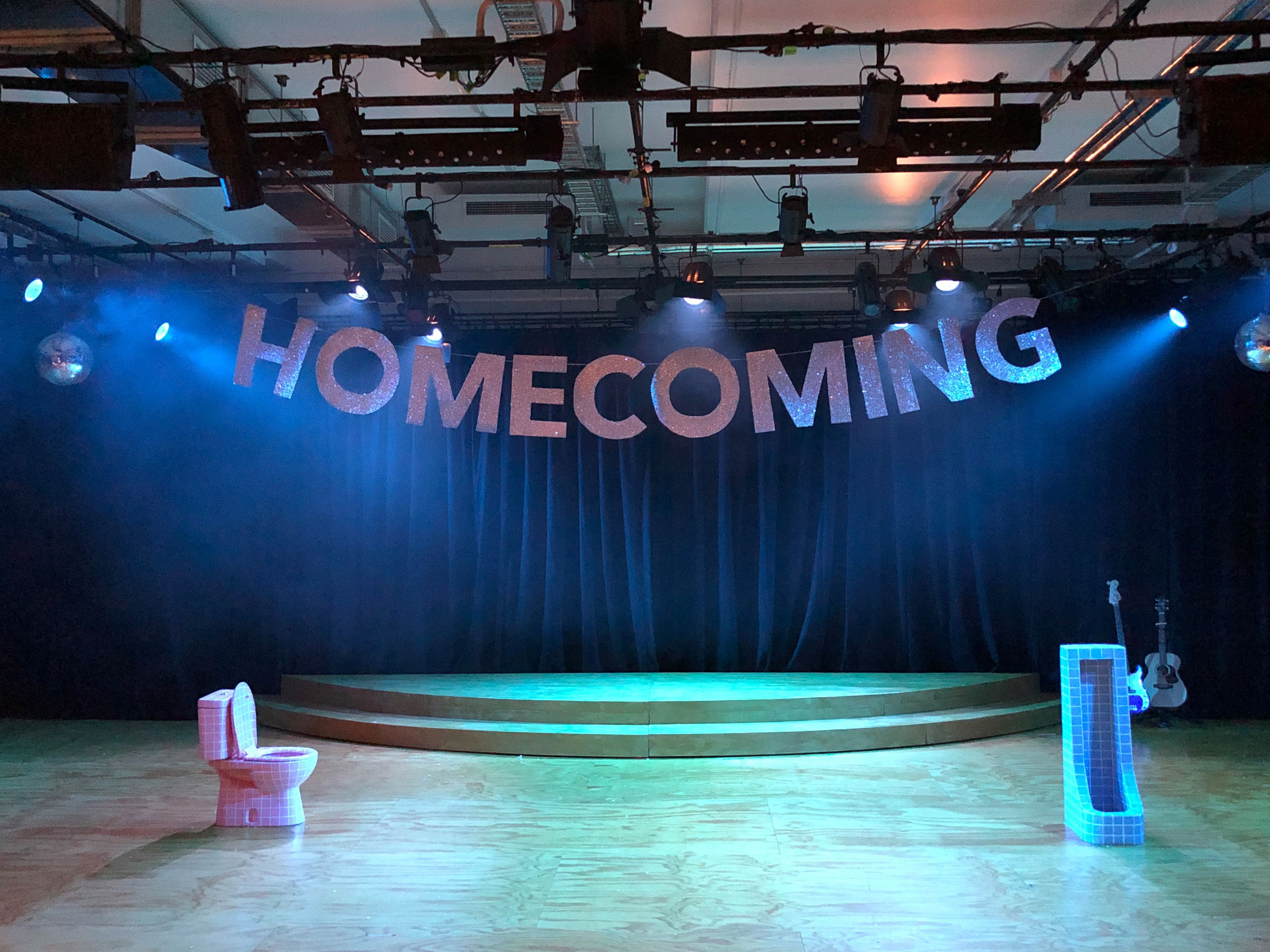 A stage setup with a glittery "HOMECOMING" banner hanging above. The stage features a wooden platform, a tiled toilet prop on the left.