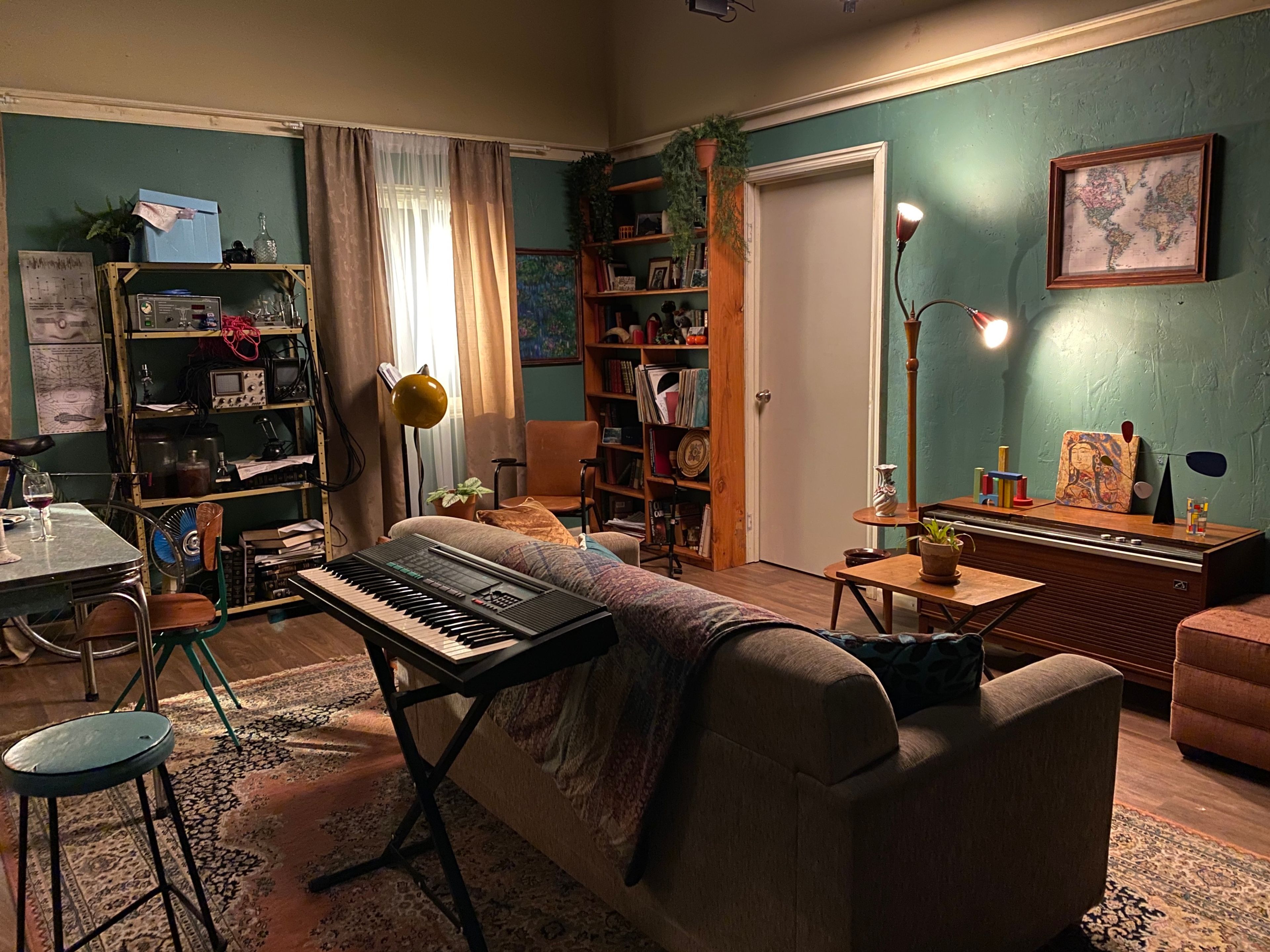 Cozy living room with teal walls, a sofa, a keyboard on a stand, bookshelves, a vintage console, and various eclectic decorations. A yellow lamp illuminates the scene