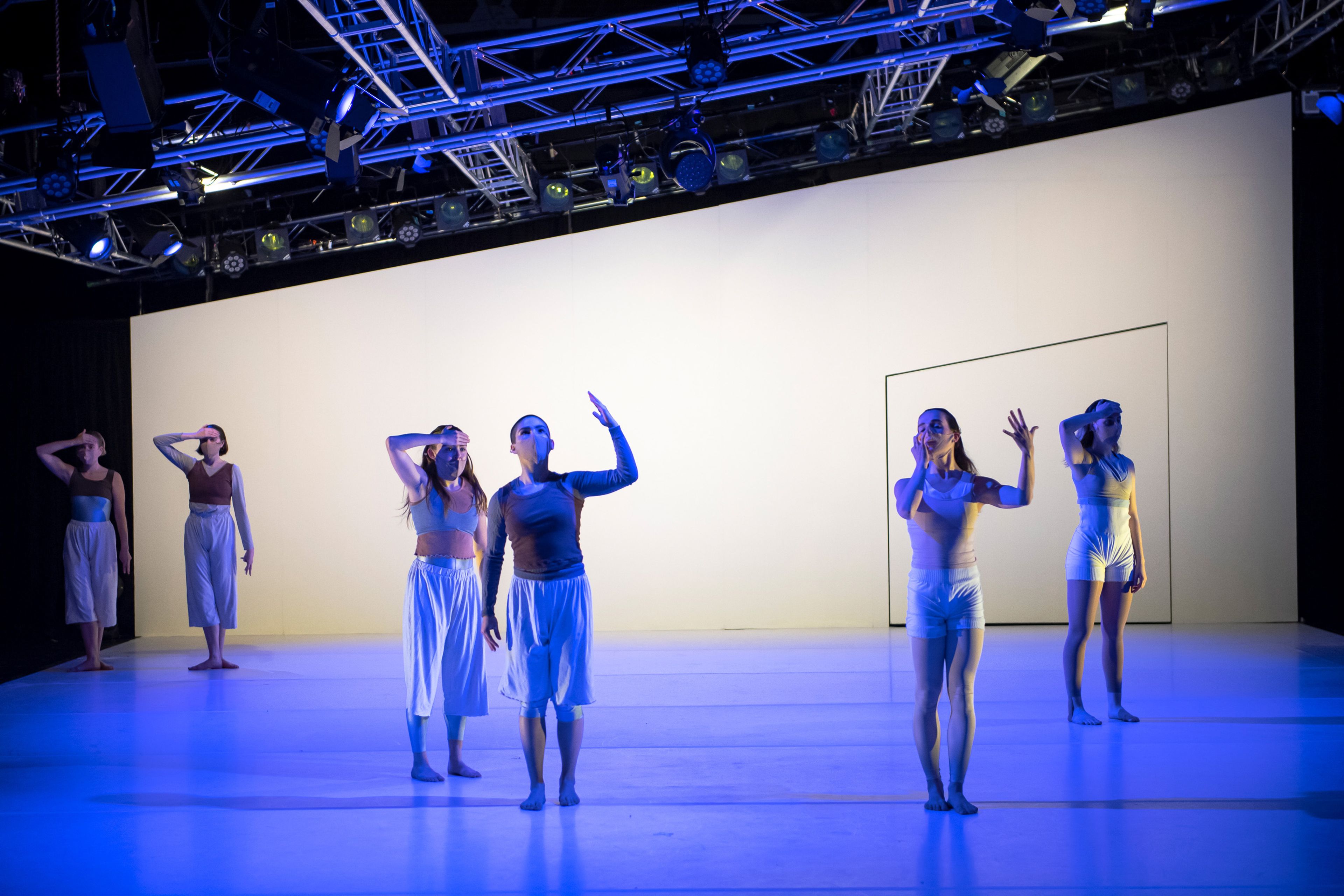 Dancers on stage, illuminated by cool blue lighting. They are arranged in a staggered formation, wearing minimalist outfits