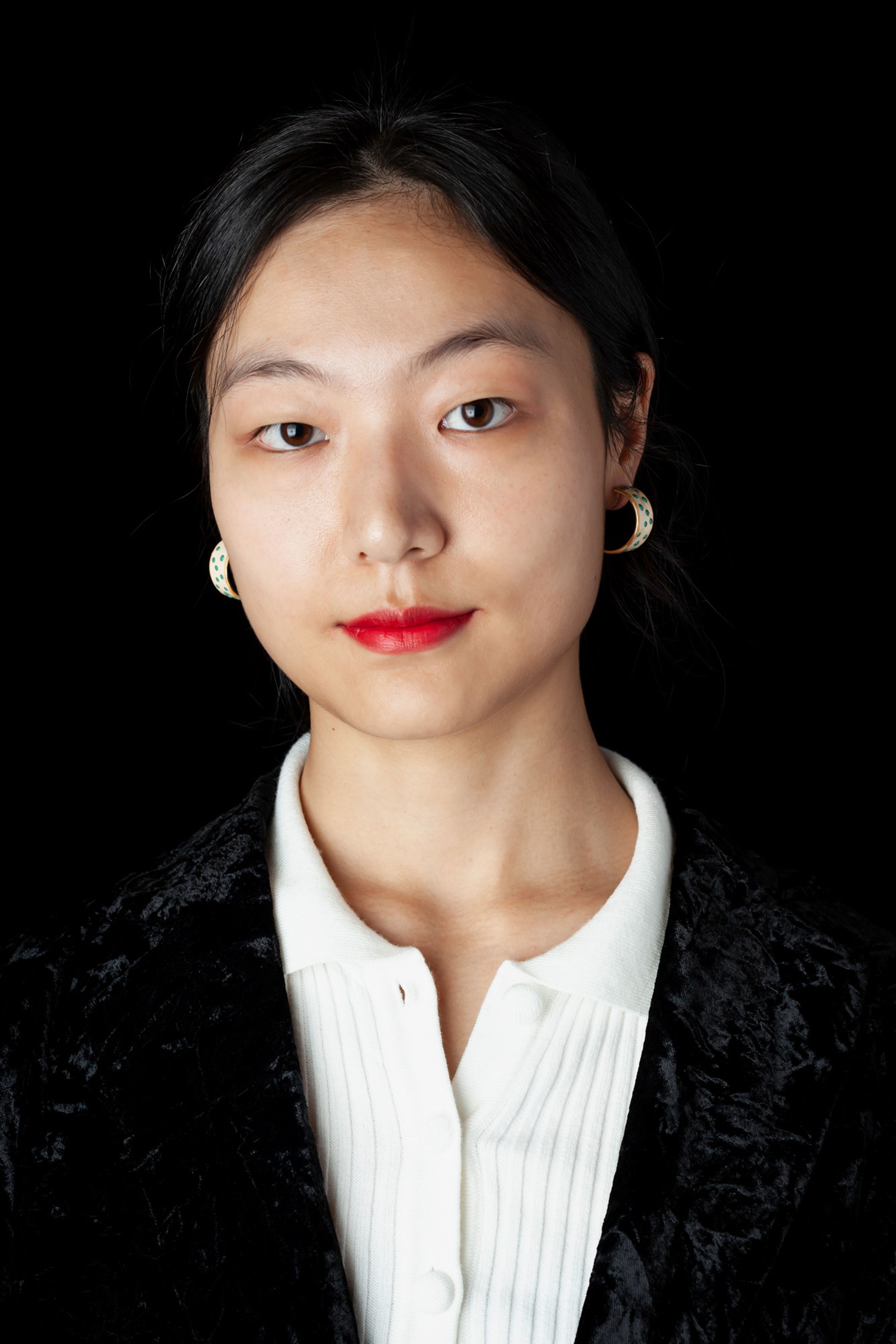 A portrait of a woman with sleek black hair, wearing statement earrings, a black textured blazer, and a white collared shirt