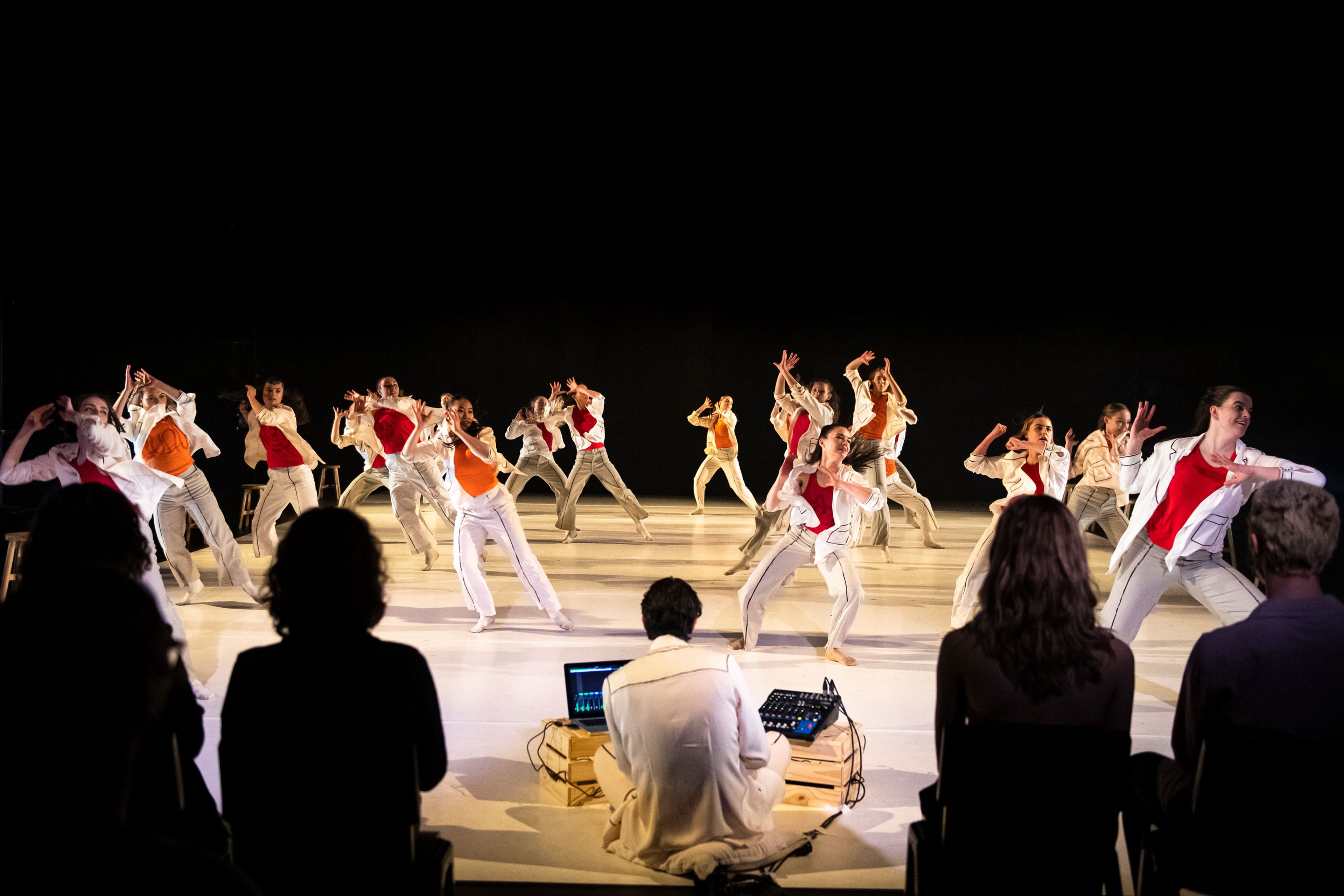 A large group of dancers in coordinated movements. The dancers are dressed in light-coloured outfits with vibrant red and orange accents. In the foreground, silhouettes of seated audience members 