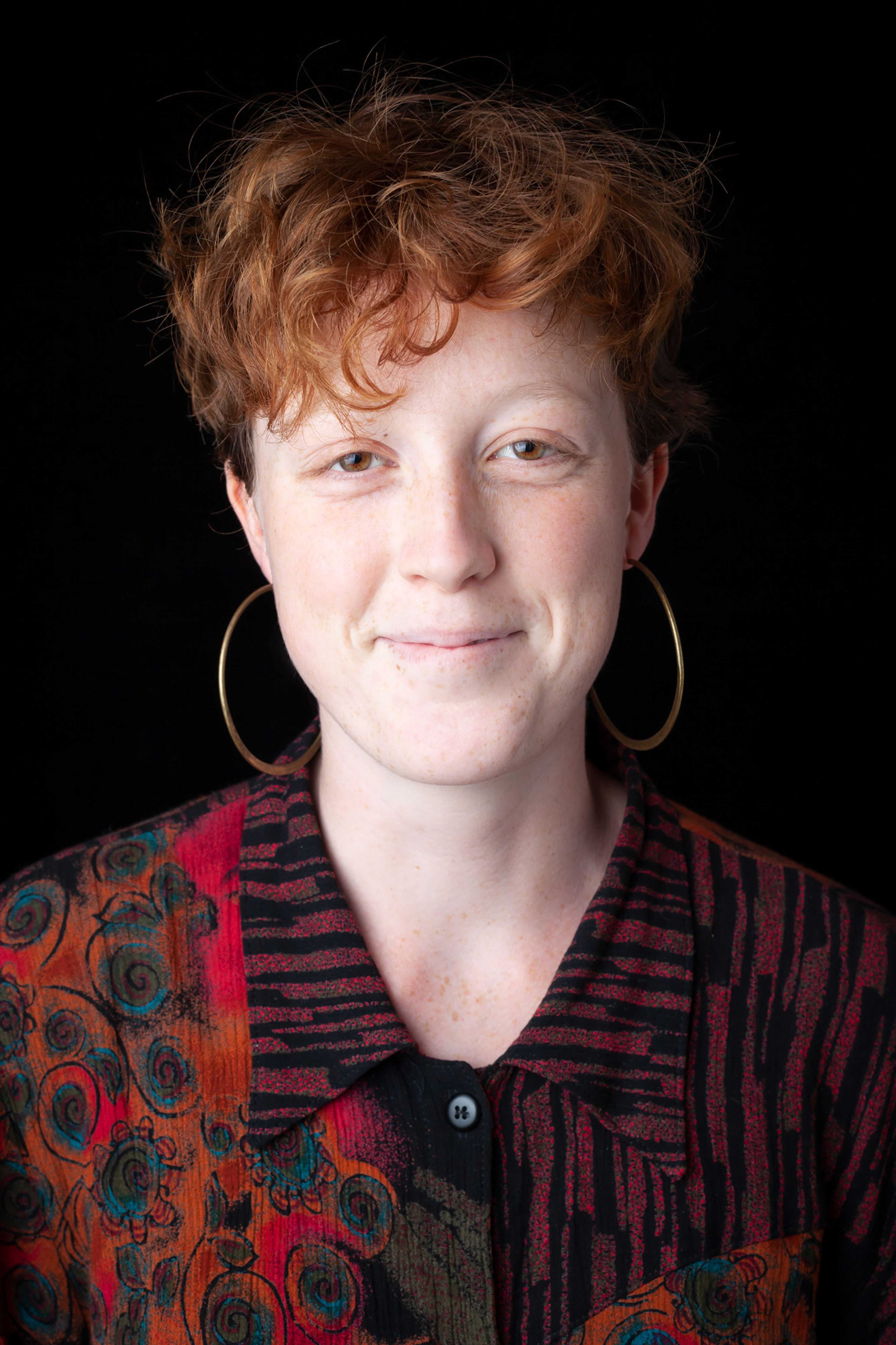 A portrait of a young person with short red hair, wearing large hoop earrings and a colorfulred and black patterned shirt