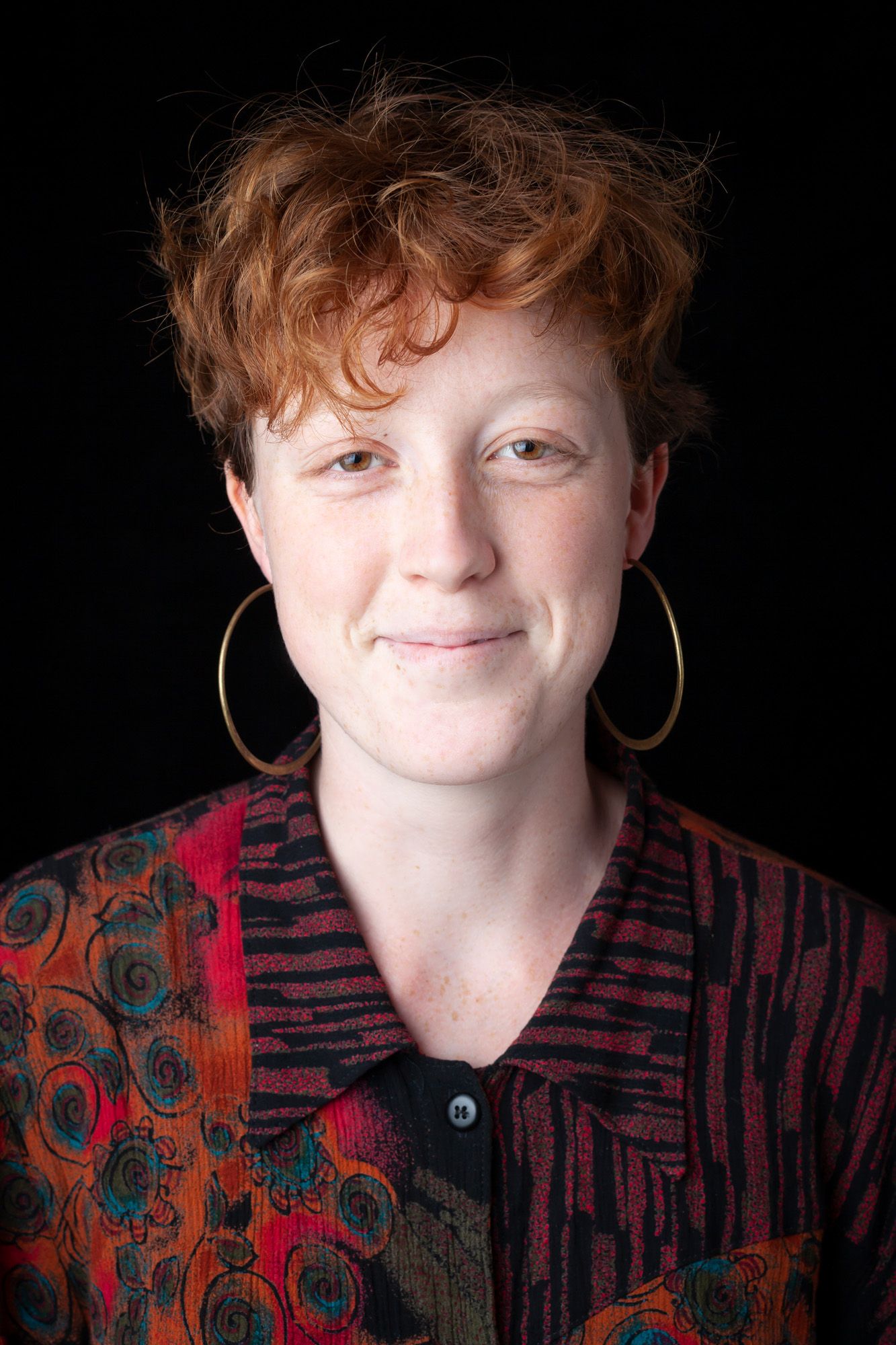 A portrait of a young person with short red hair, wearing large hoop earrings and a colorfulred and black patterned shirt