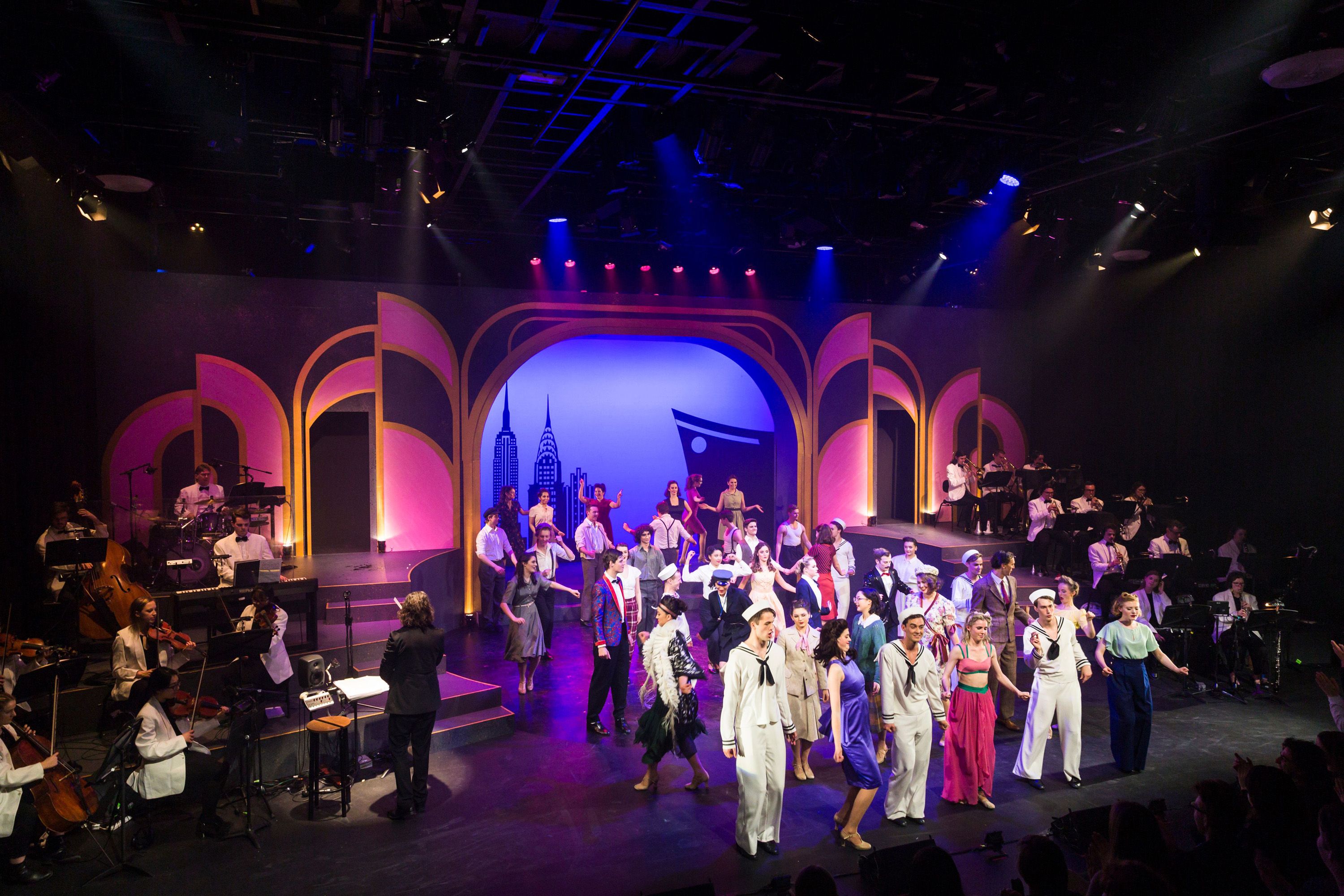 Stage performance featuring a large ensemble cast in vibrant costumes, including sailors, dancers, and musicians. The backdrop displays an art deco-style set with a silhouette of a city skyline