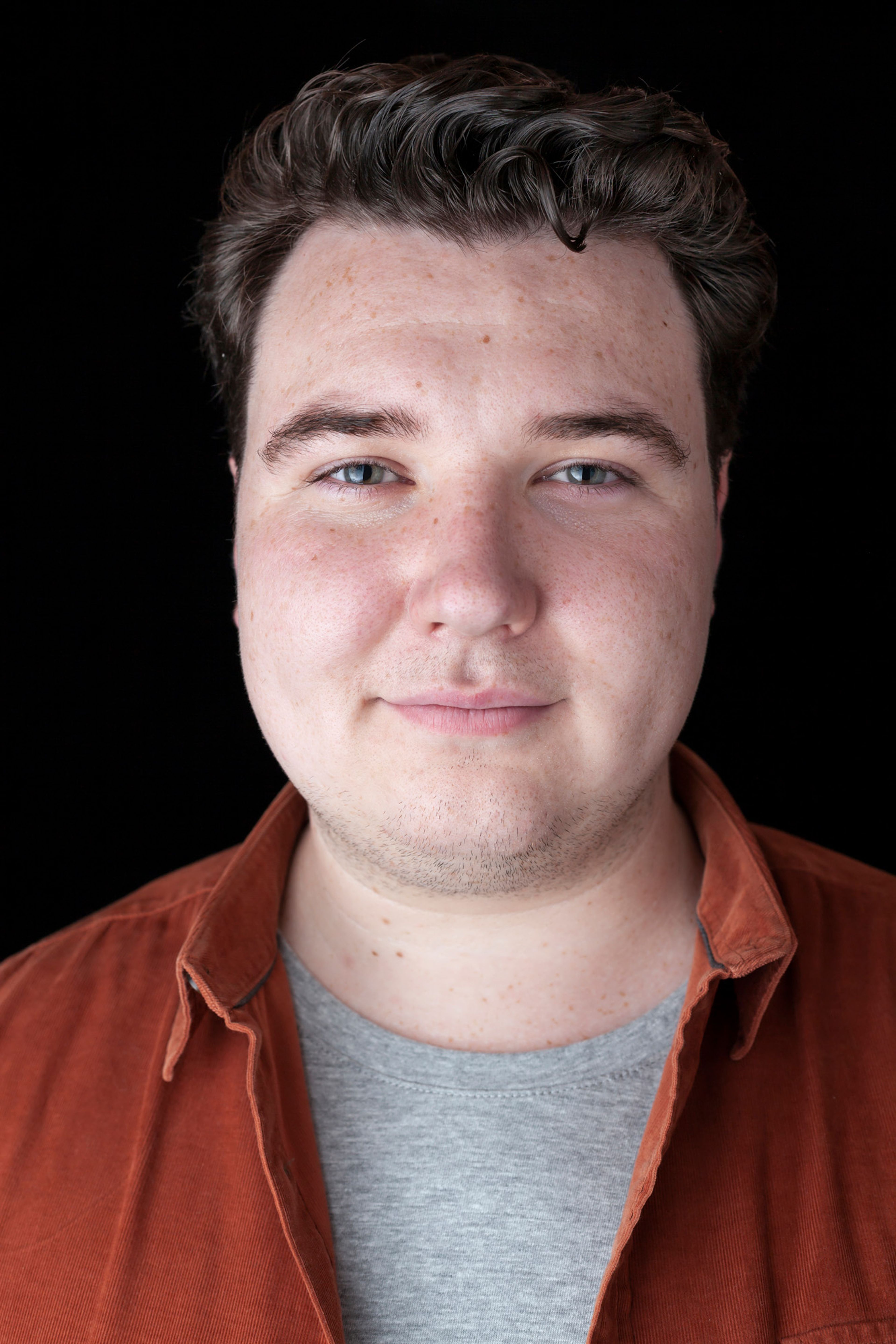 A portrait of a man with short, dark hair, wearing a rust-colored shirt over a gray t-shirt. He has a neutral expression