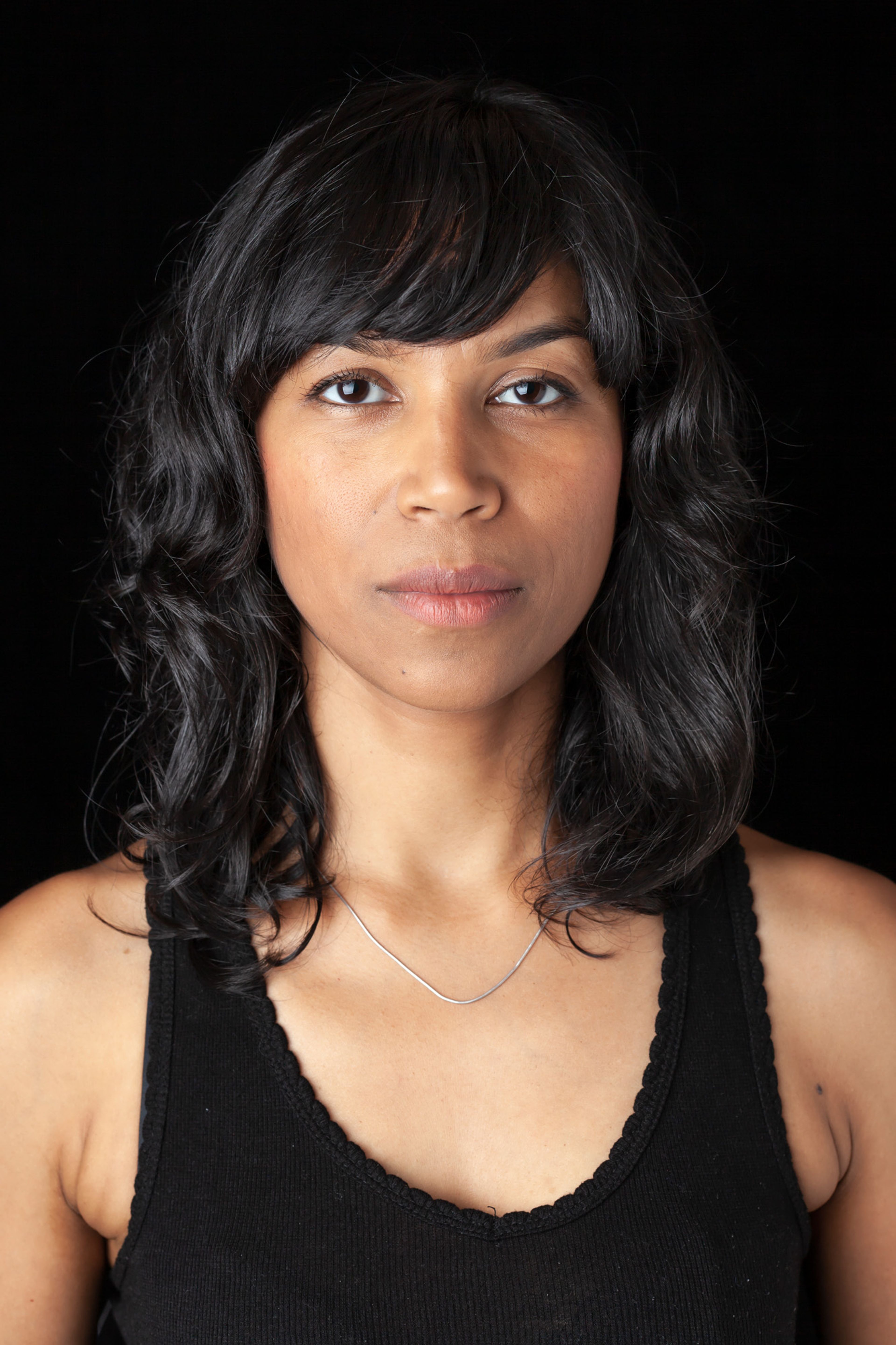 Portrait of a woman with shoulder-length wavy black hair, wearing a black tank top and a thin silver necklace