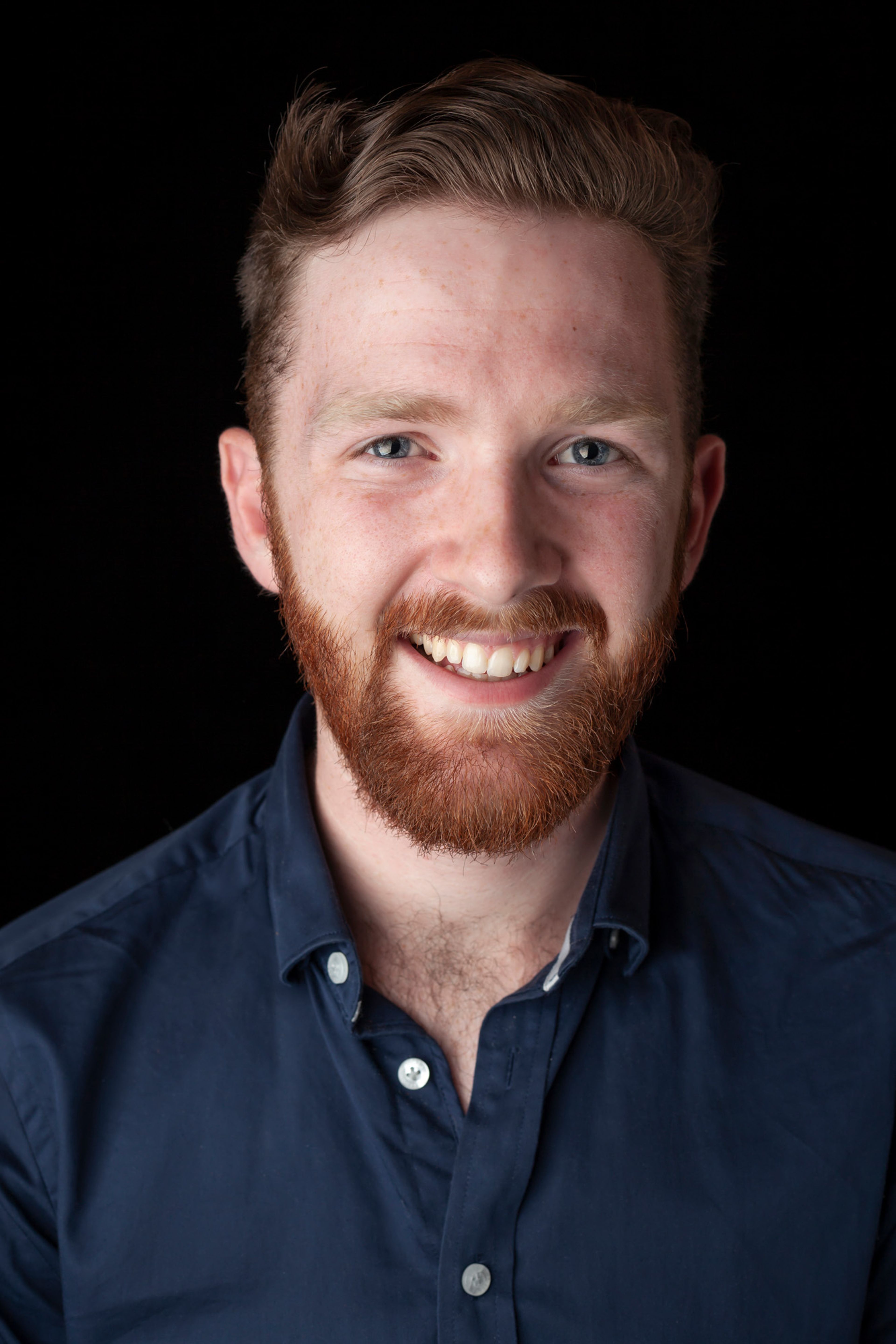 A portrait of a smiling man with a red beard and short, neatly styled hair. He is wearing a navy blue button-down shirt