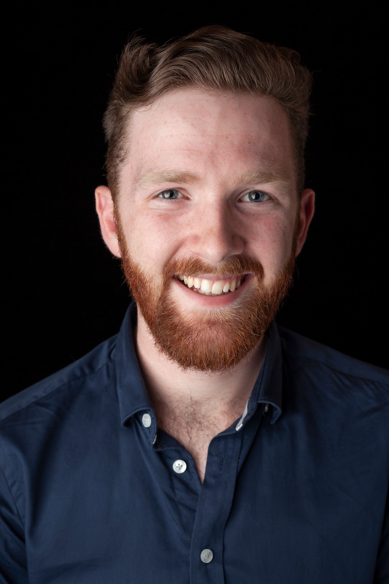 A portrait of a smiling man with a red beard and short, neatly styled hair. He is wearing a navy blue button-down shirt