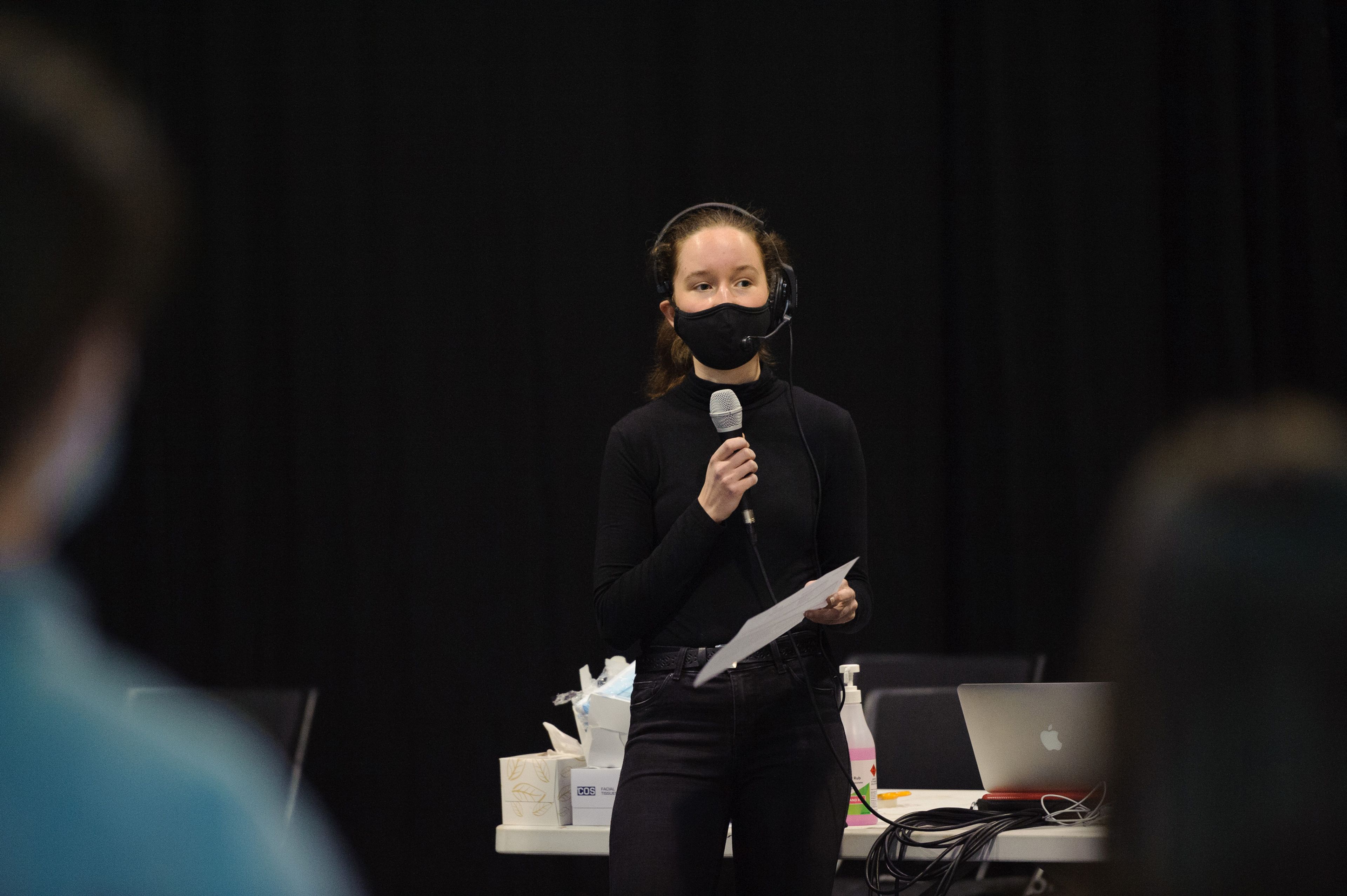 A person wearing a black mask and a headset. They hold a microphone in one hand and a piece of paper in the other, speaking to the production team