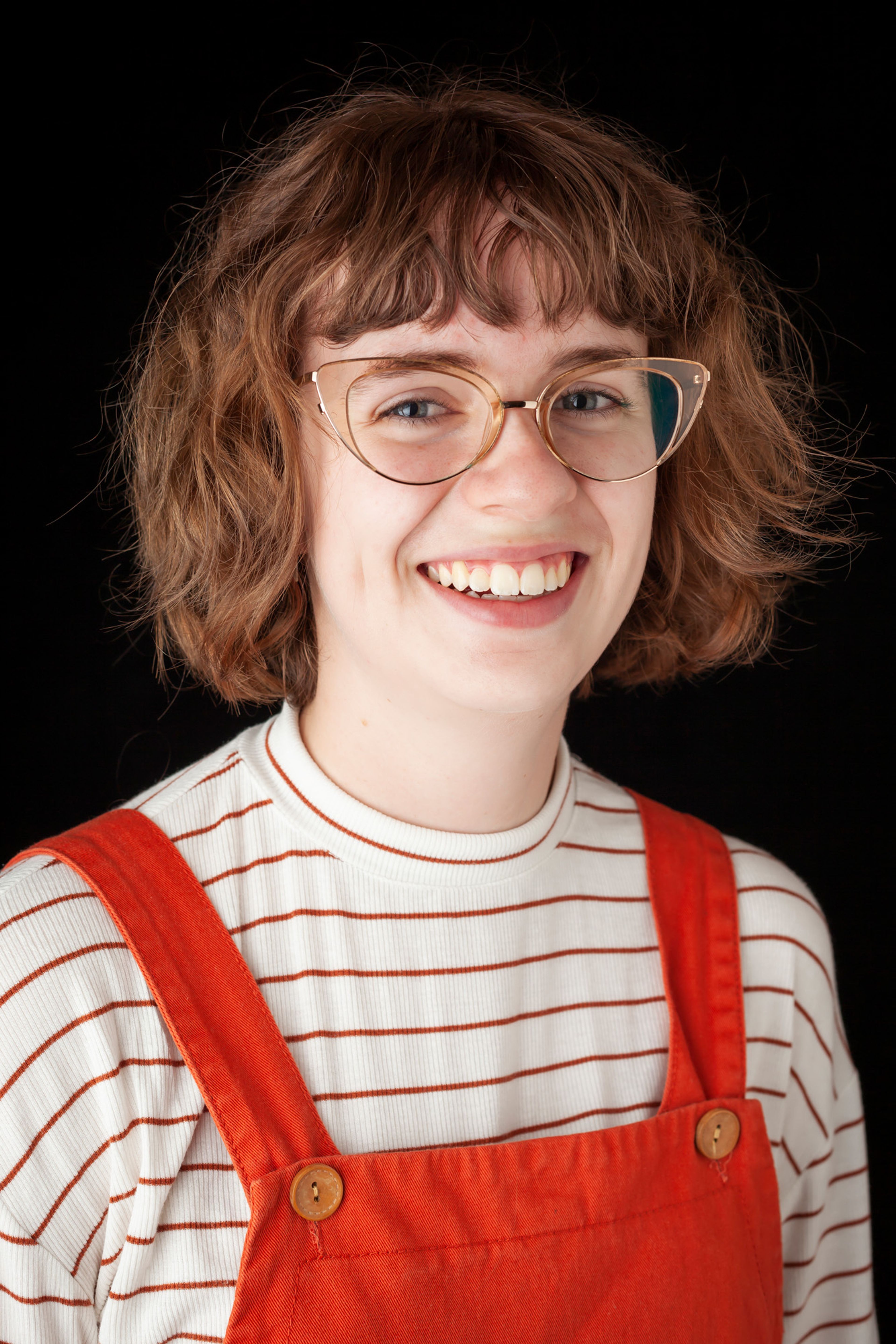 A smiling individual with short, wavy hair wearing round glasses and a striped shirt paired with orange overalls