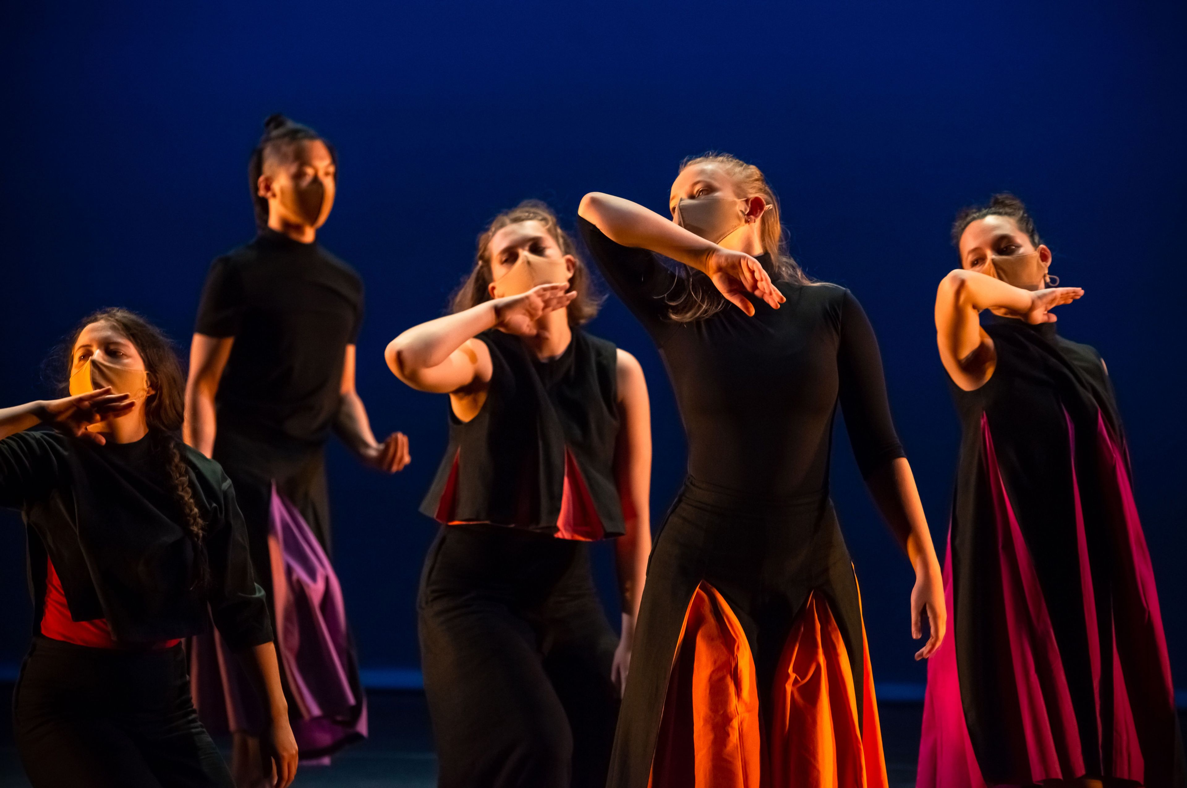 Contemporary dance performance featuring five dancers wearing black outfits with vibrant red, orange, and purple accents. They perform synchronised movements with their arms raised, against a deep blue backdrop