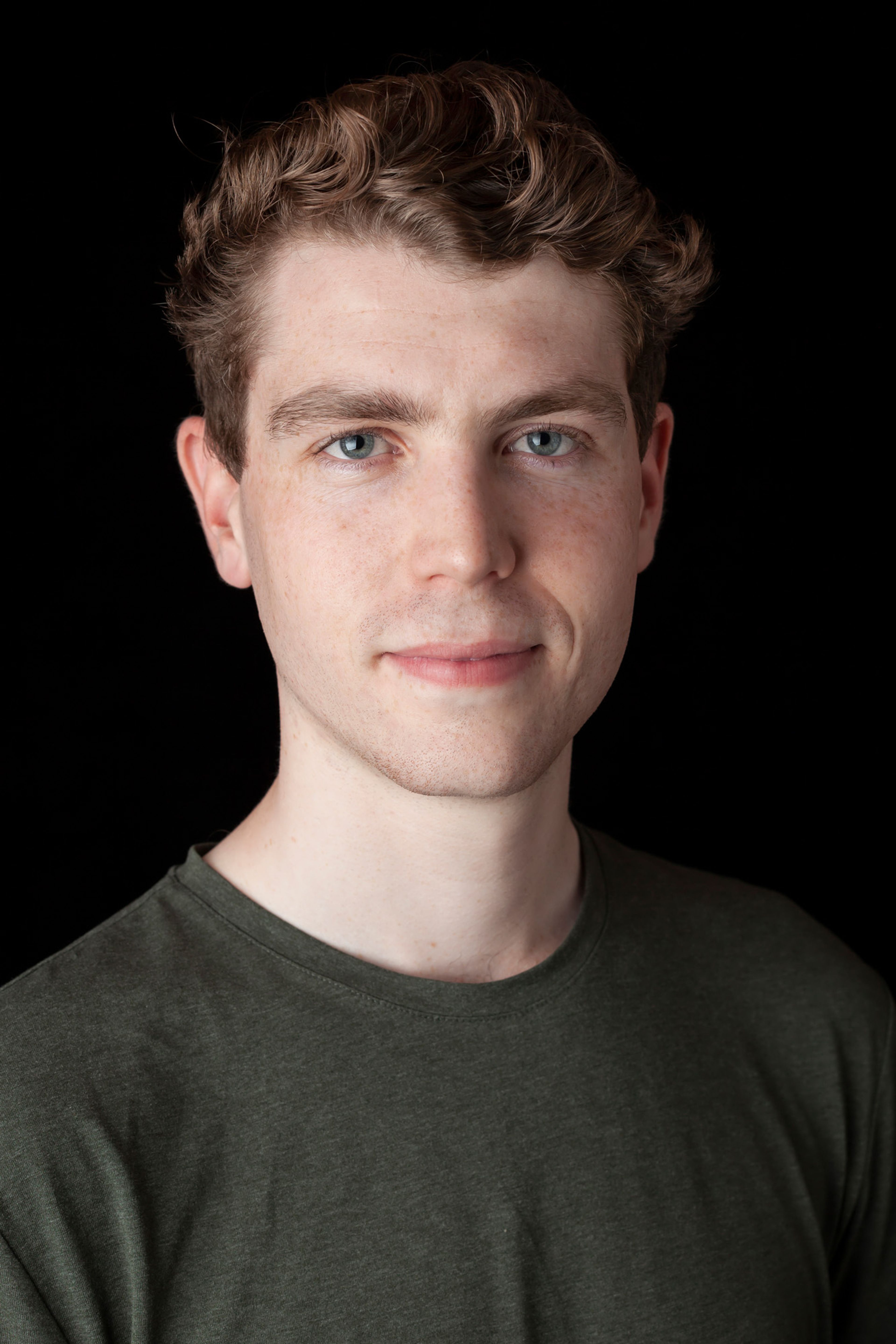 A portrait of a man with short, wavy hair, light skin, and blue eyes, wearing a dark green t-shirt