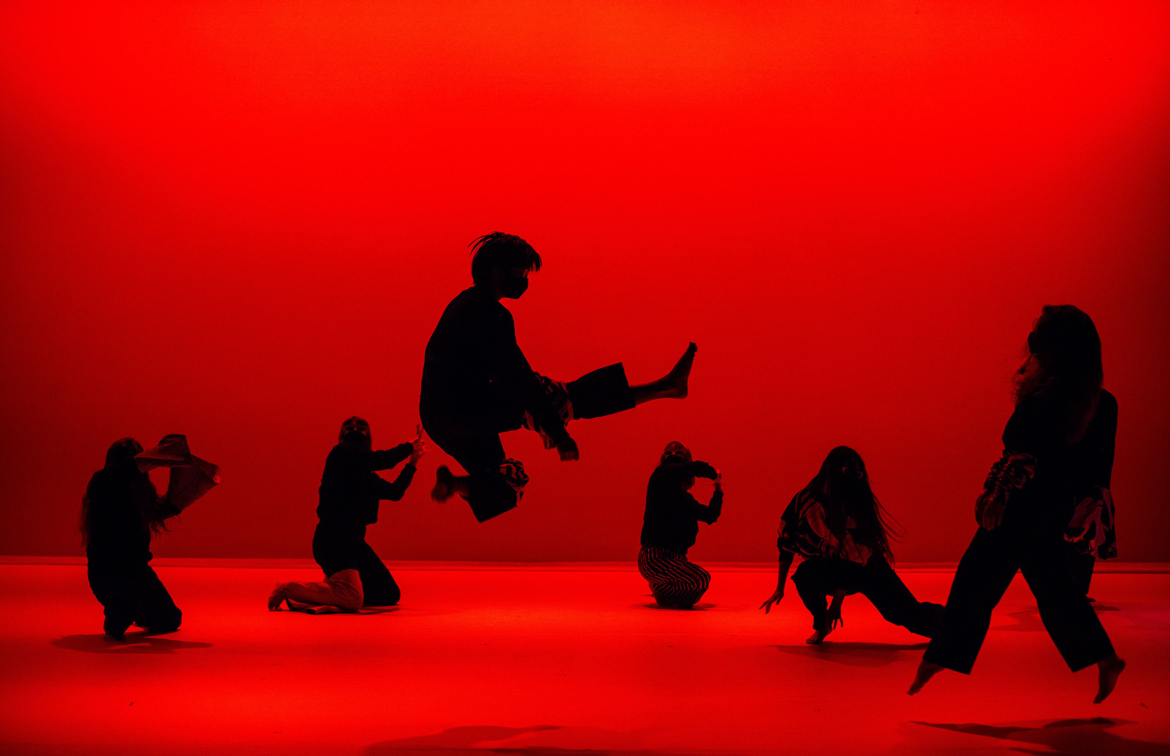 Dramatic performance against a vivid red background, silhouetted figures are dynamically posed, with one figure mid-air in an impressive leap.