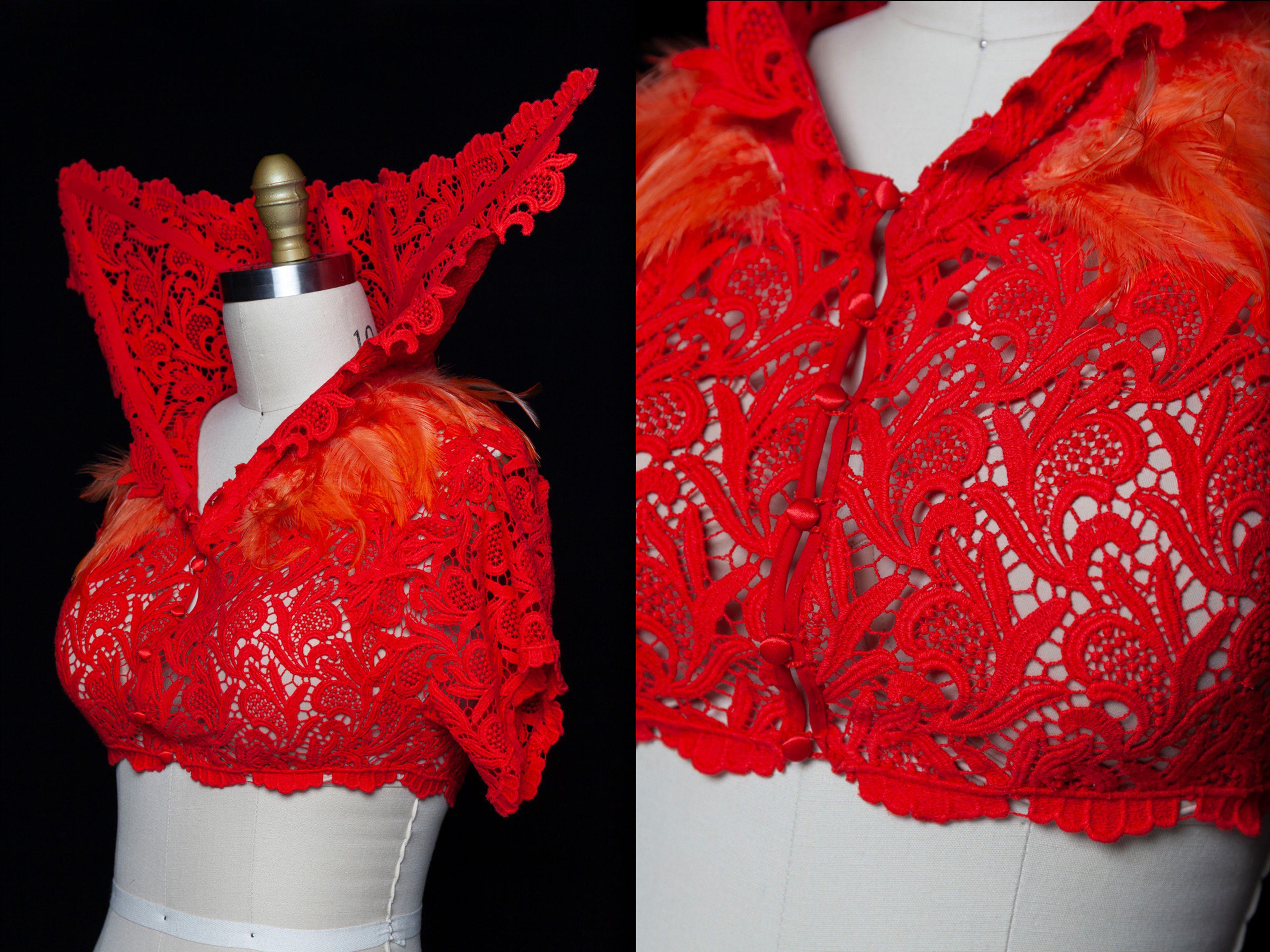 A vibrant red lace garment displayed on a mannequin, featuring intricate floral patterns and a dramatic raised collar. The design is accented with orange feathers along the shoulders