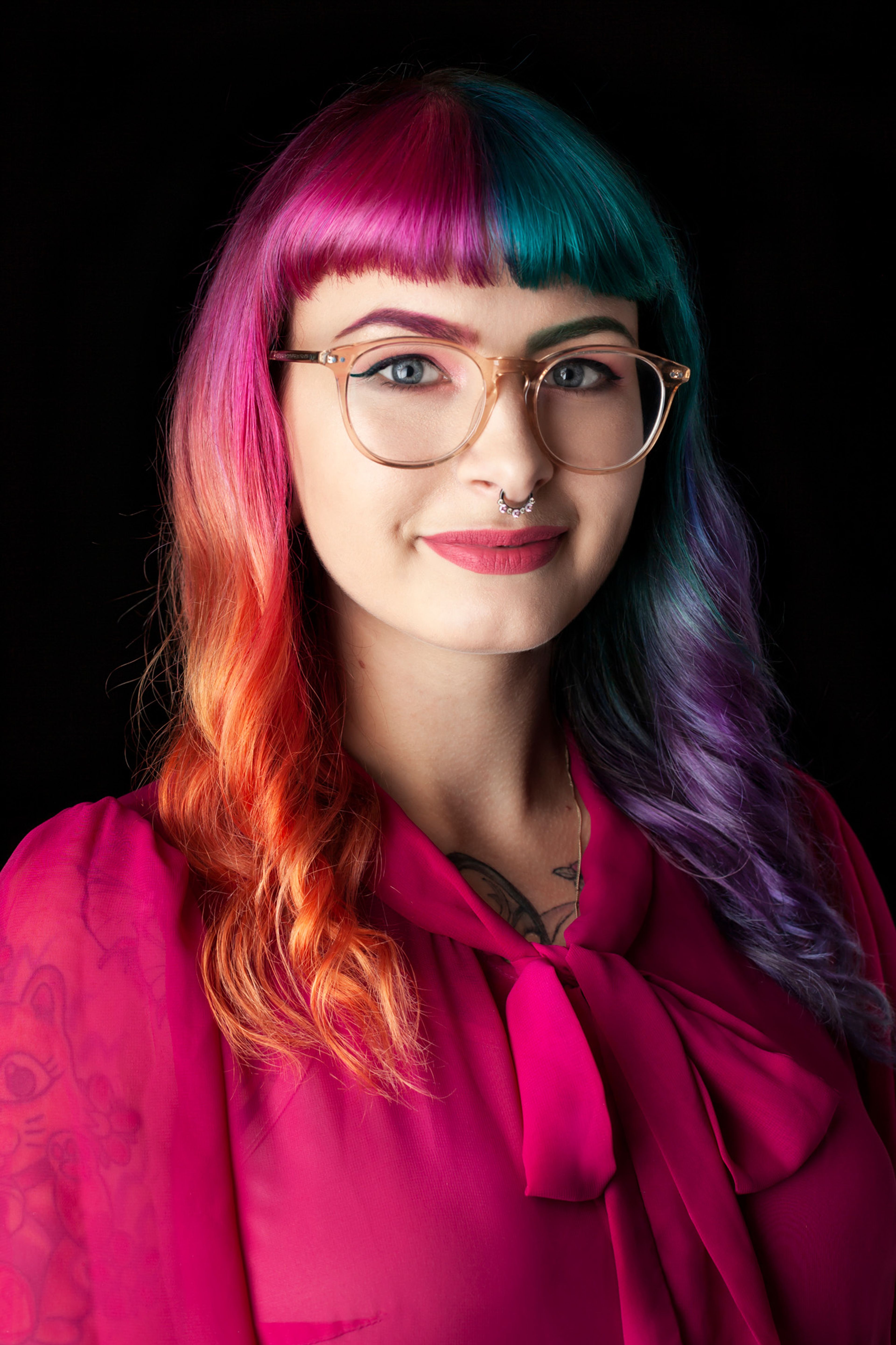 A portrait of a woman with brightly colored hair, split into vibrant pink and teal, styled with bangs. She wears glasses, and a magenta blouse with a bow detail.