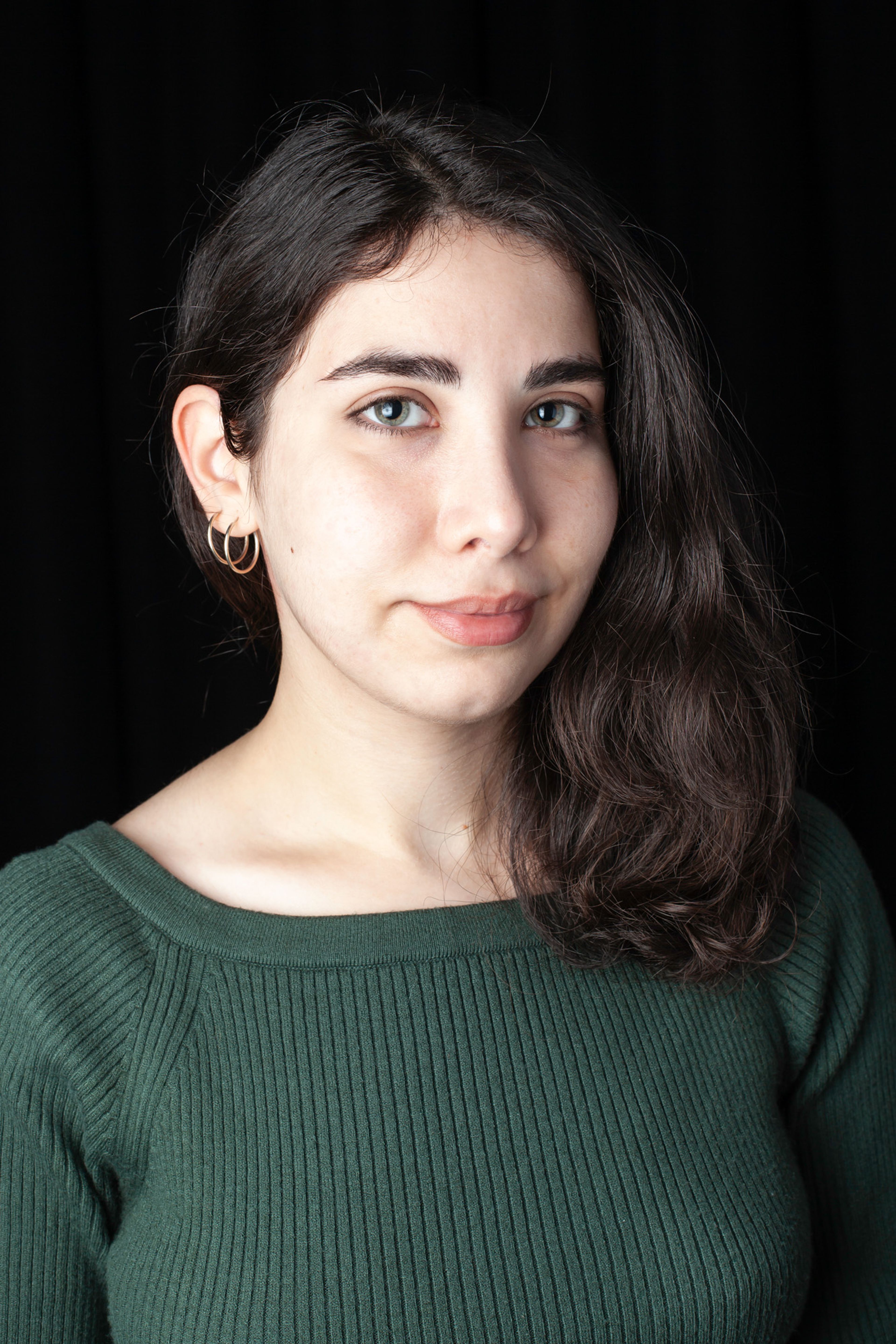 Portrait of a person with wavy dark hair, wearing a green ribbed top and subtle gold hoop earrings