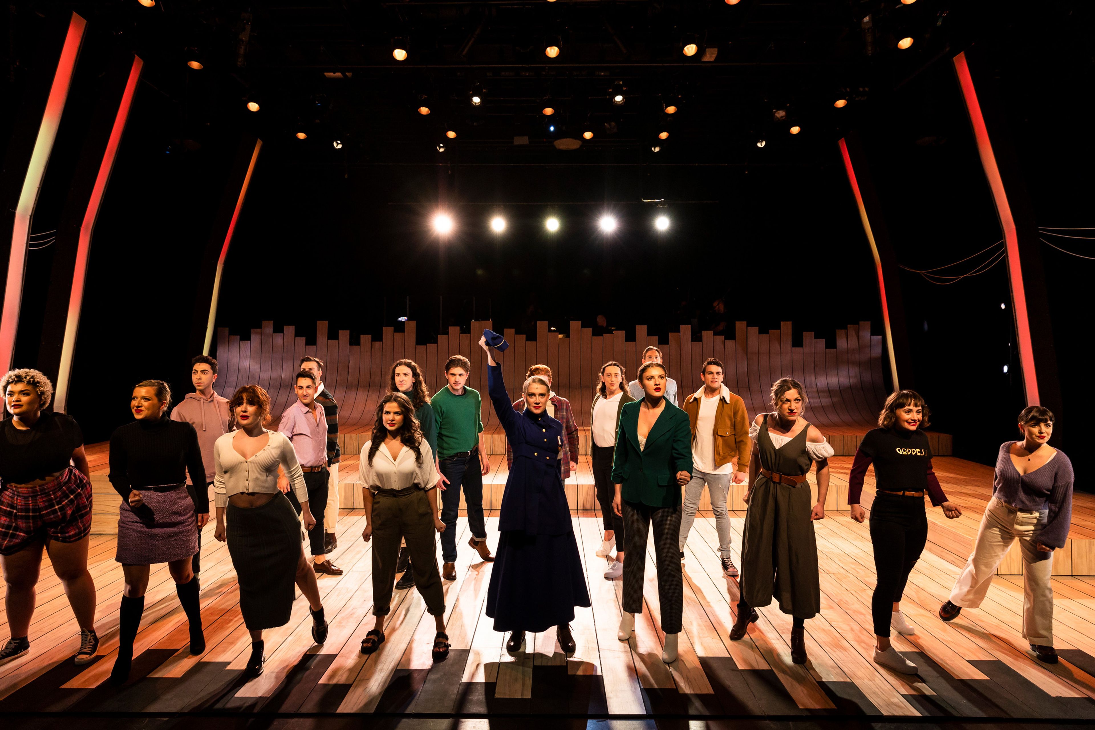 Ensemble of performers on a brightly lit wooden stage, framed by colourful vertical light strips. The group stands in a powerful formation