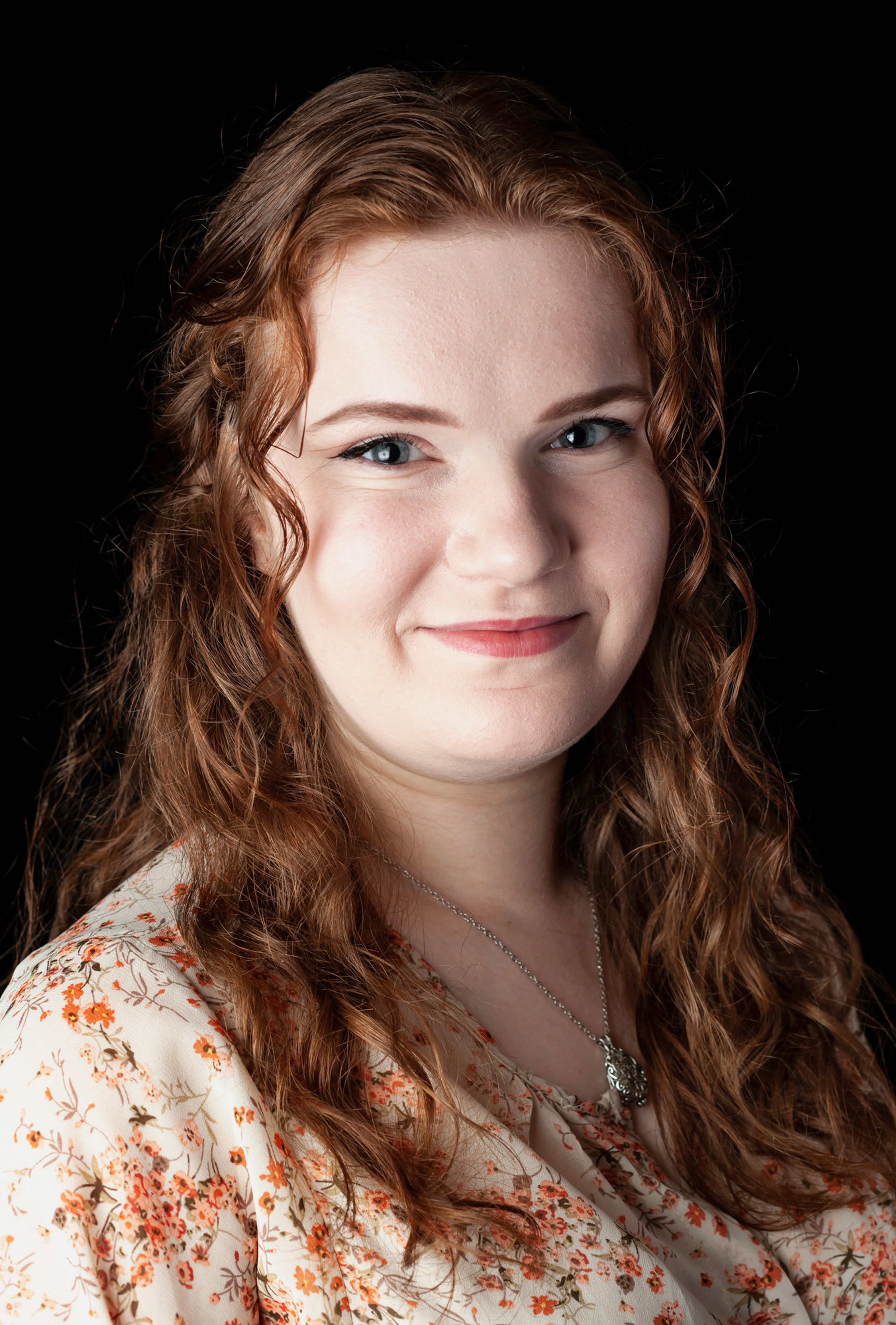 The image is a professional headshot of a person with long, wavy red hair, wearing a floral-patterned top and a pendant necklace