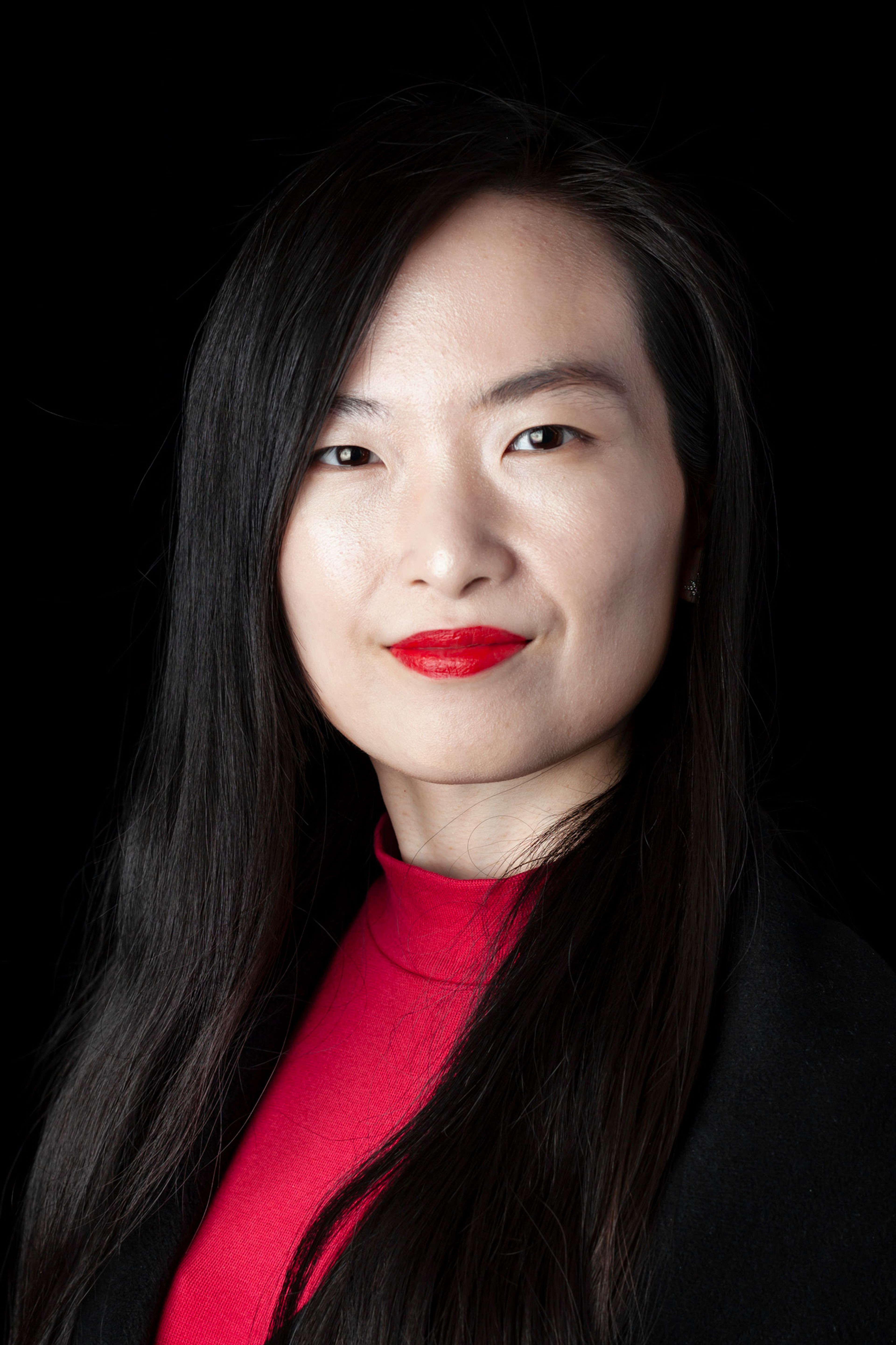 Headshot of a woman with long black hair, styled sleek and straight, wearing a vibrant red top and bold red lipstick