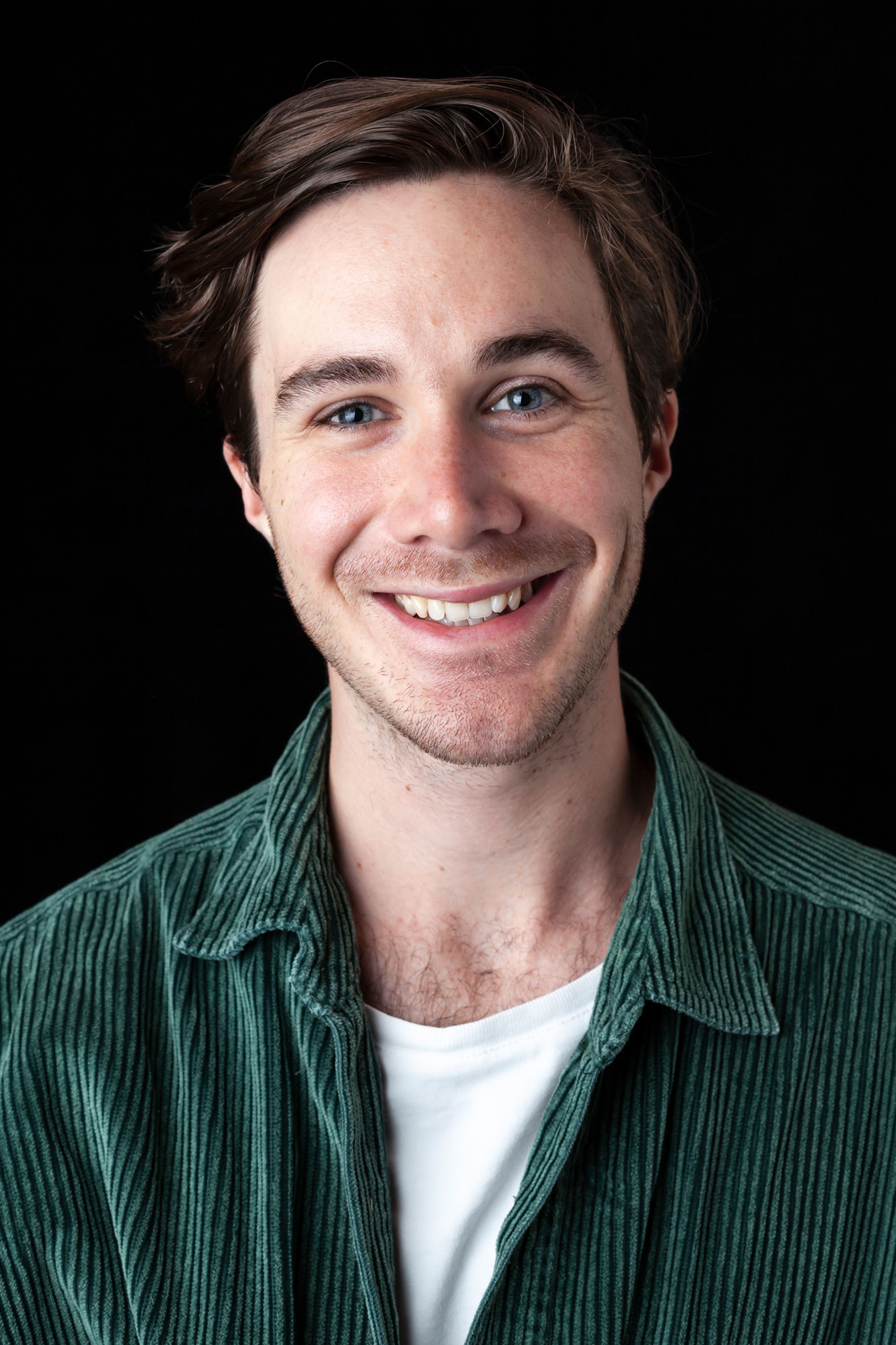 A portrait of a smiling person with short, wavy hair, wearing a green corduroy shirt over a white top