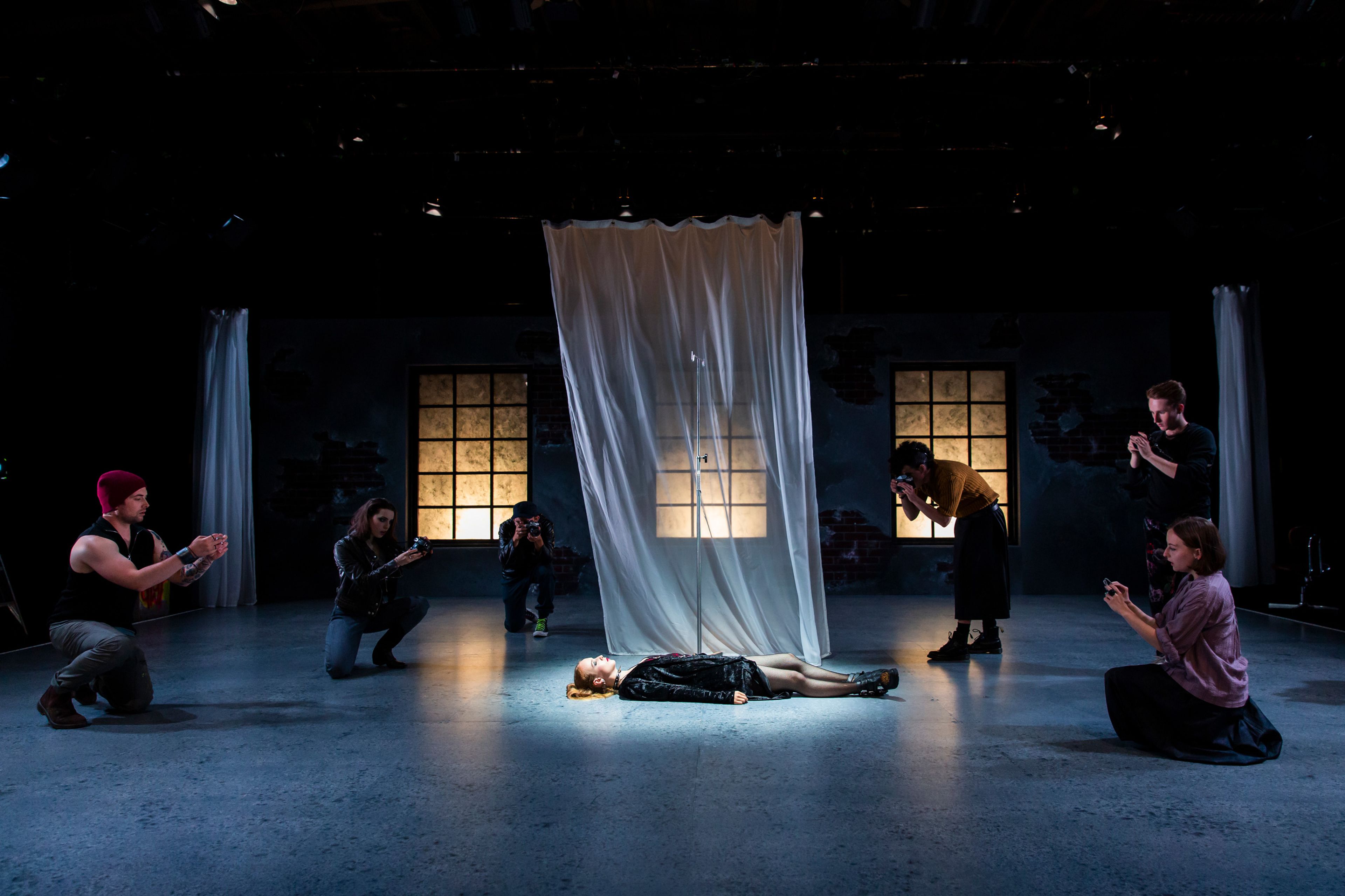 A production photograph a person lying on stage under a spotlight surrounded by other performers crouching and photographing. The stage is dimly lit, with illuminated windows in the background