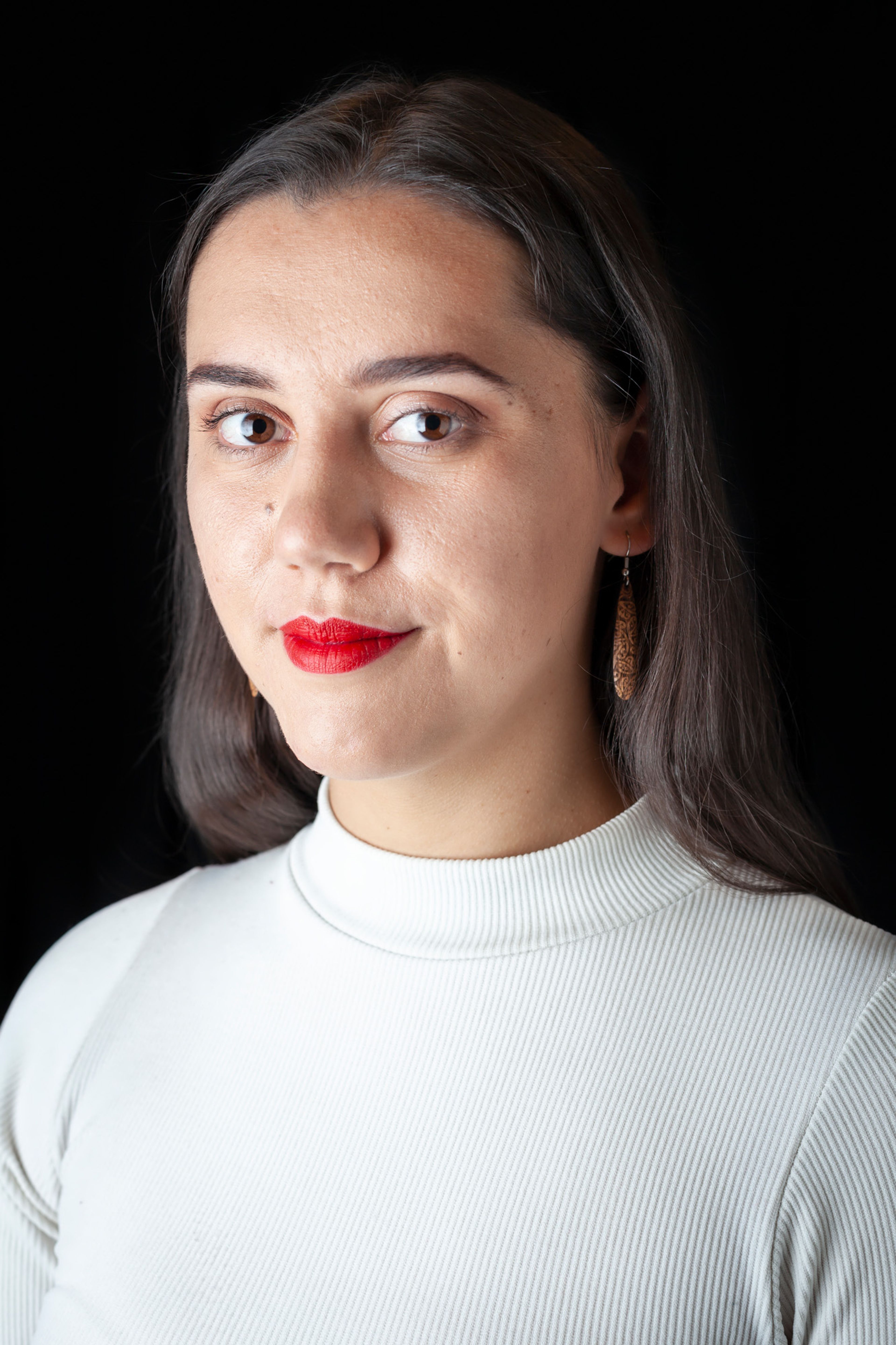 A portrait of a woman with long dark hair, wearing a white ribbed high-neck top and wooden drop earrings. She has bright red lipstick