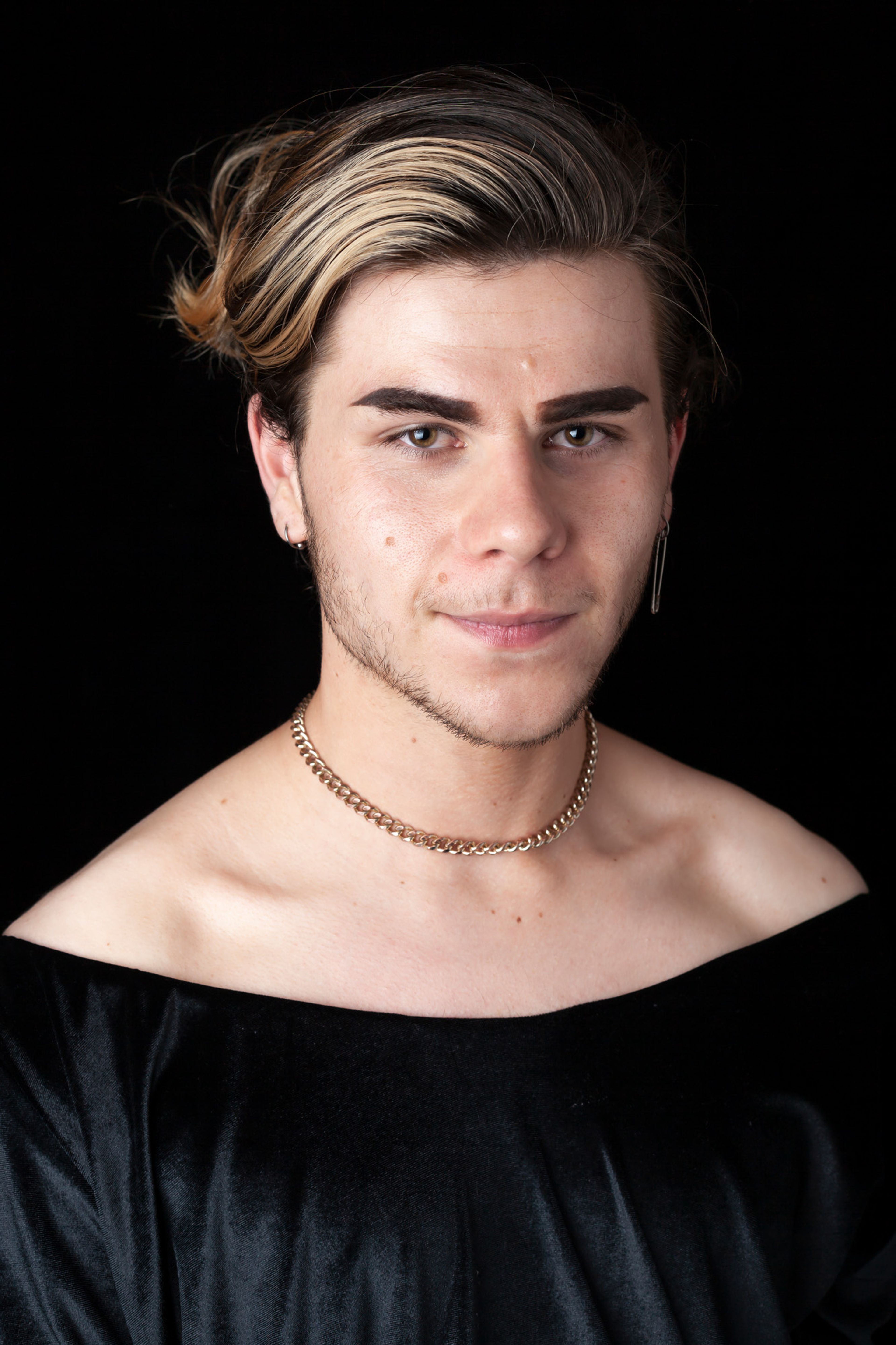 A portrait of a person with styled, highlighted hair, wearing a black off-shoulder top, a gold chain necklace, and a single drop earring. They have bold eyebrows and a confident expression.