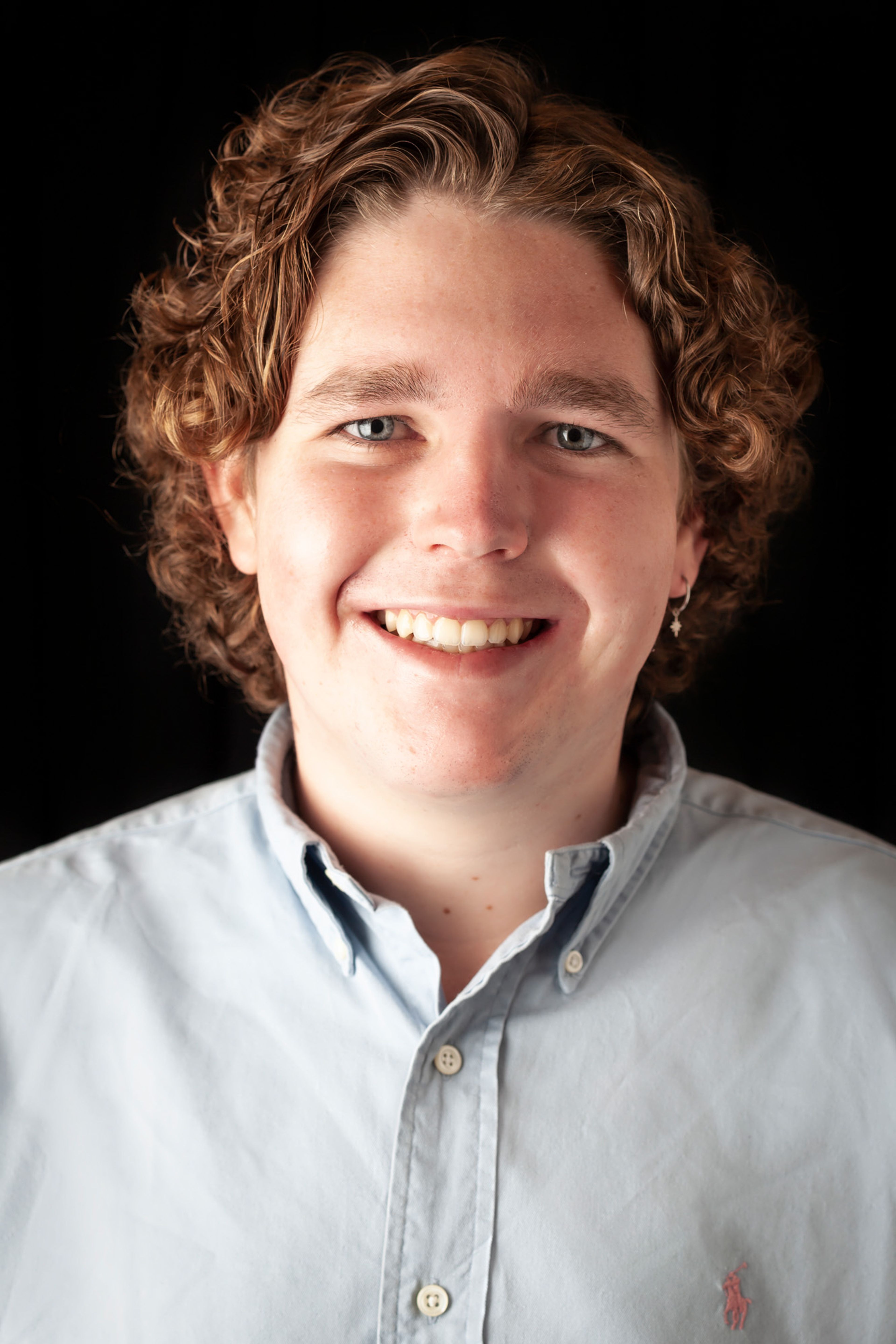 A portrait of a person with curly light brown hair, wearing a light blue button-up shirt, smiling