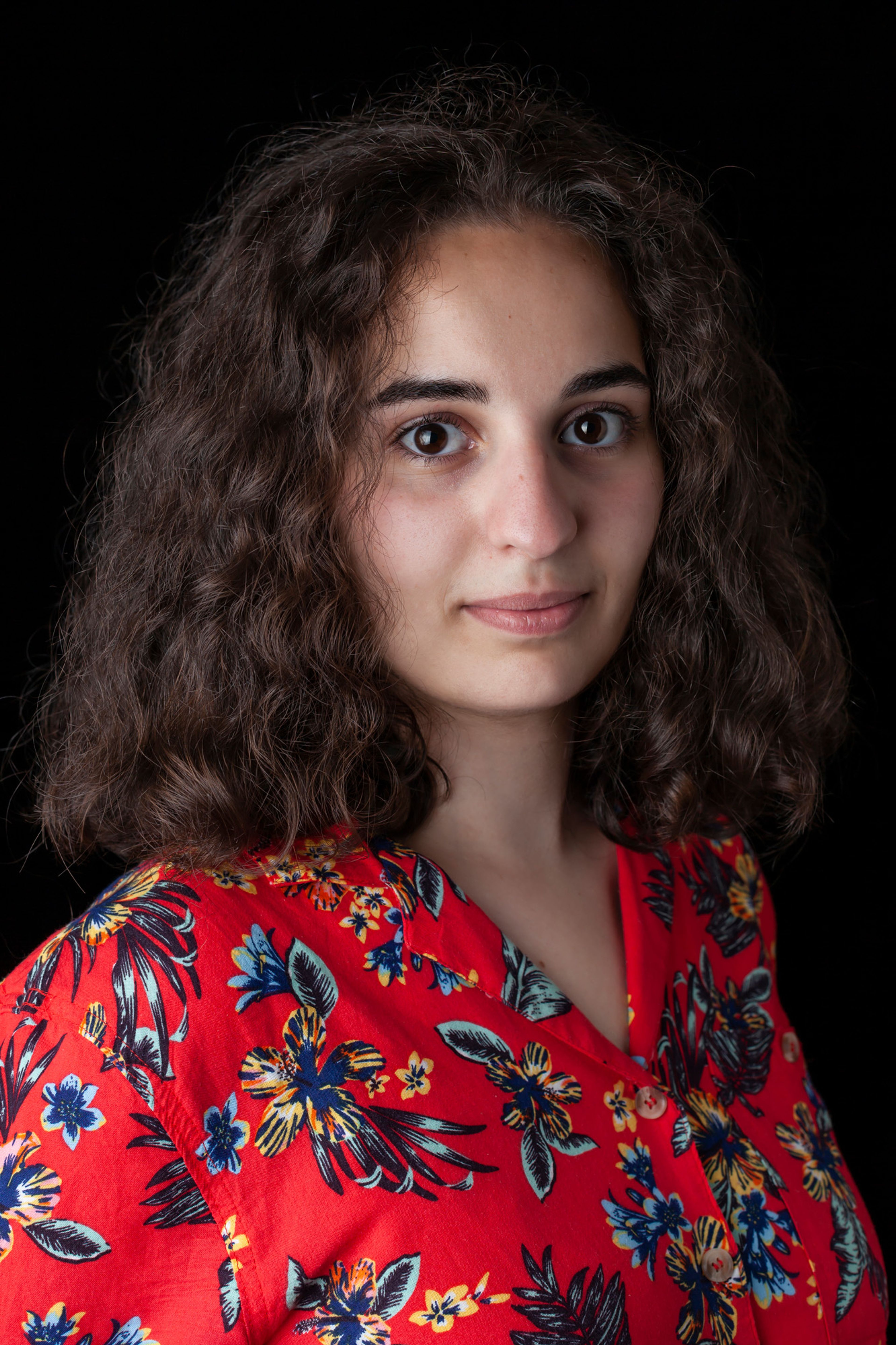 Portrait of a person with shoulder-length curly brown hair and dark eyes, wearing a vibrant red shirt with a floral pattern