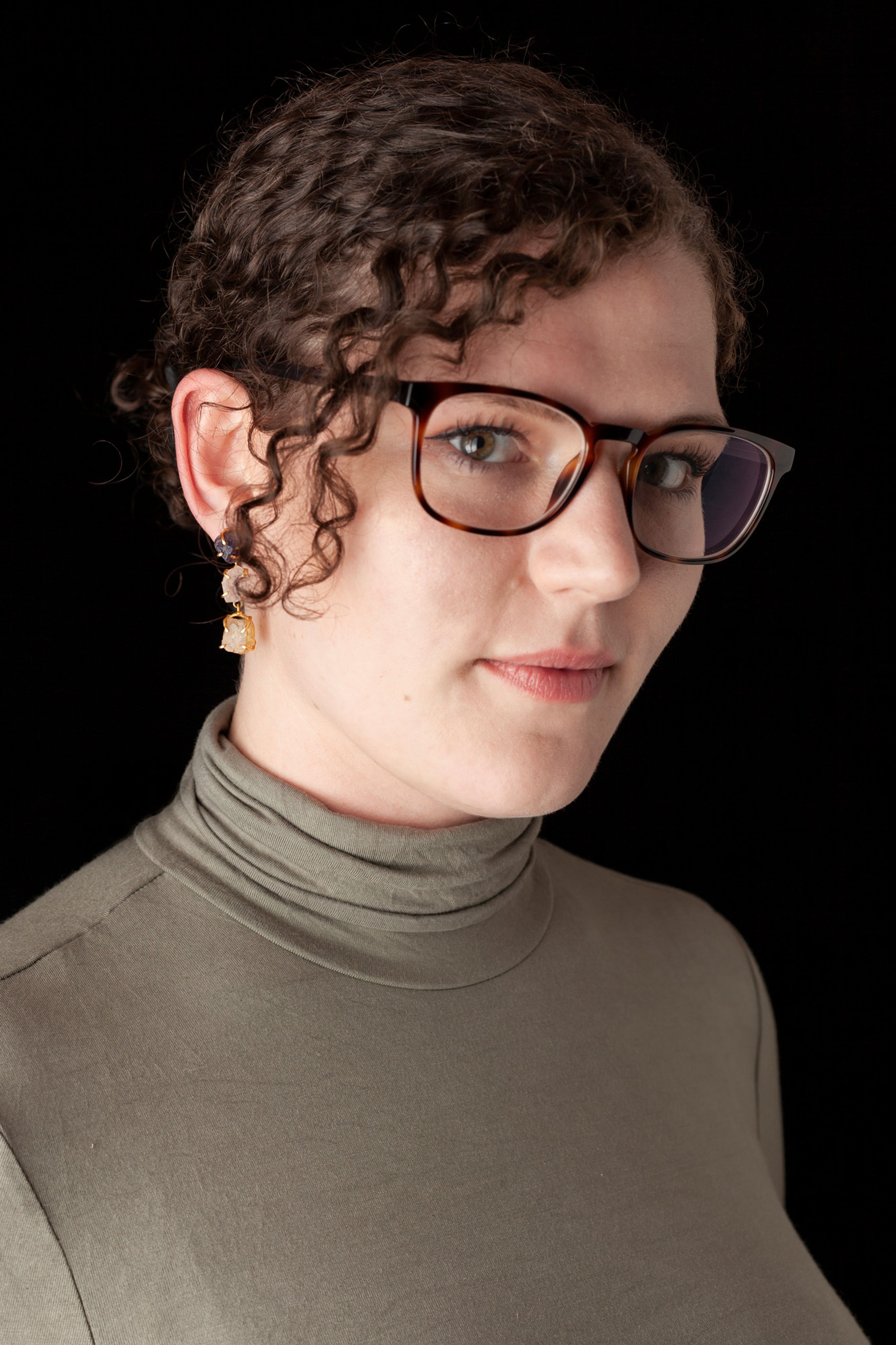 A portrait of a young woman with curly hair, wearing tortoiseshell glasses, drop earrings, and an olive-green turtleneck