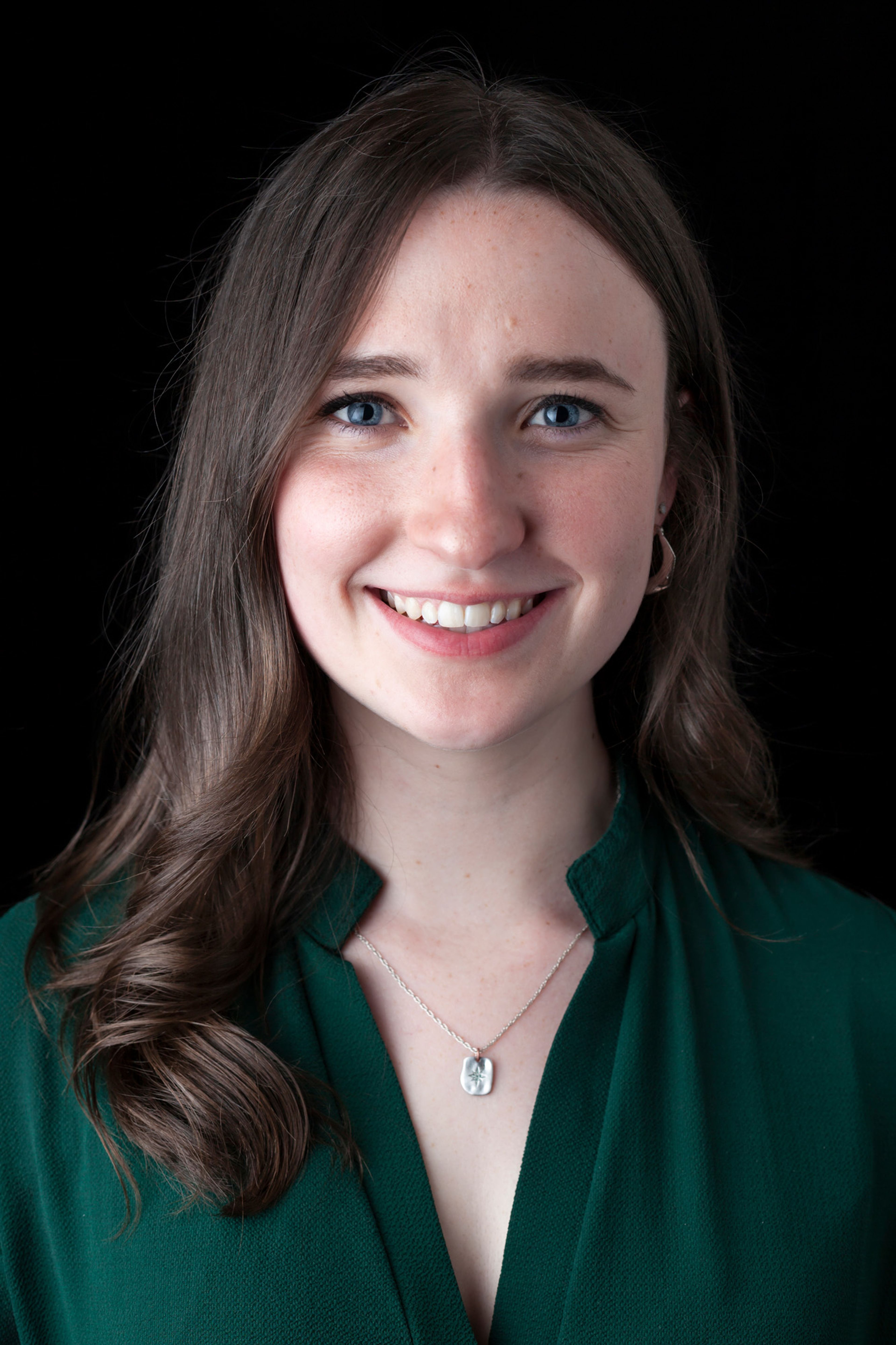 Portrait of a smiling person with long, wavy brown hair and blue eyes. They are wearing a green blouse