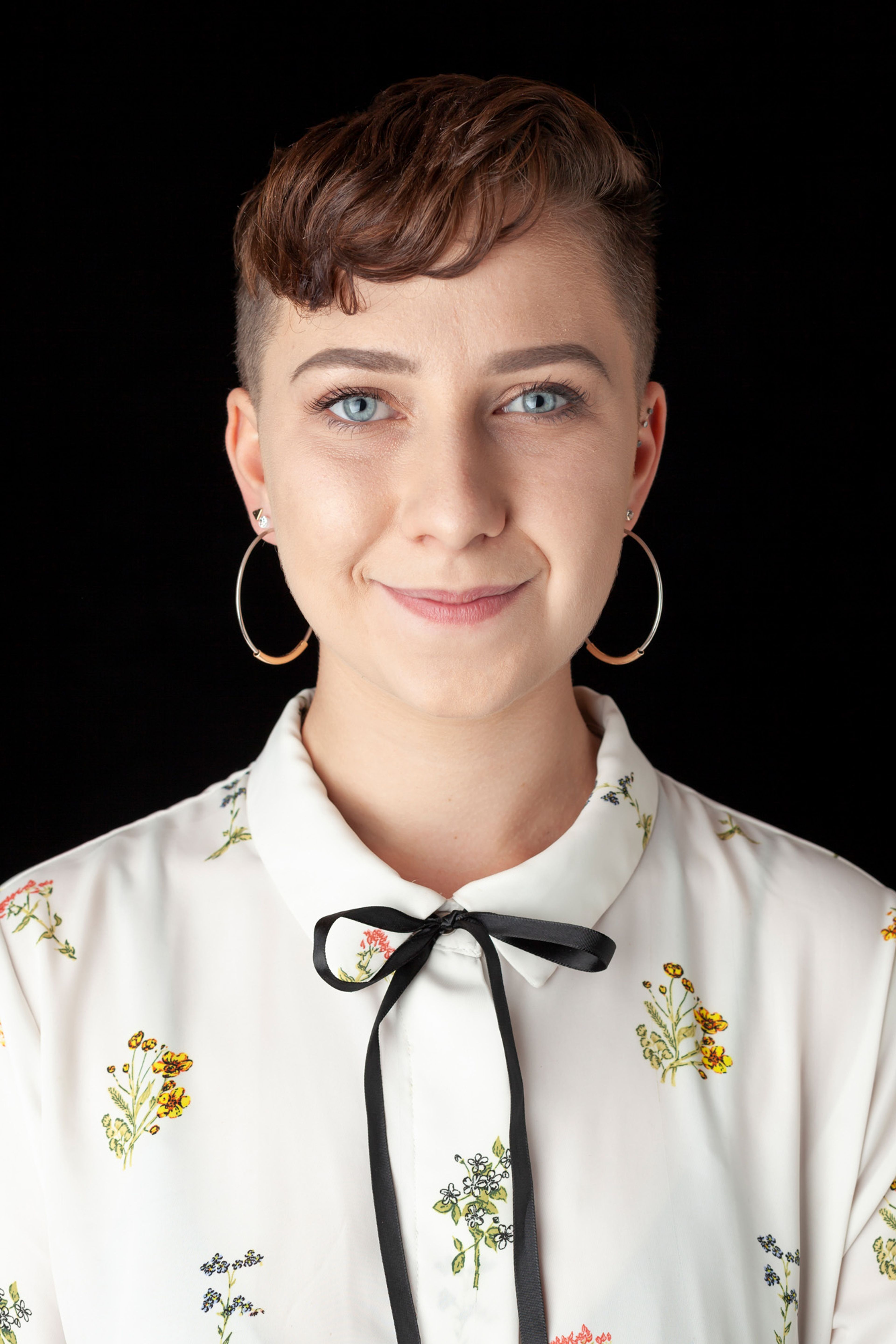 A portrait of a woman with short hair, wearing hoop earrings and a white blouse with floral patterns and a black ribbon tie