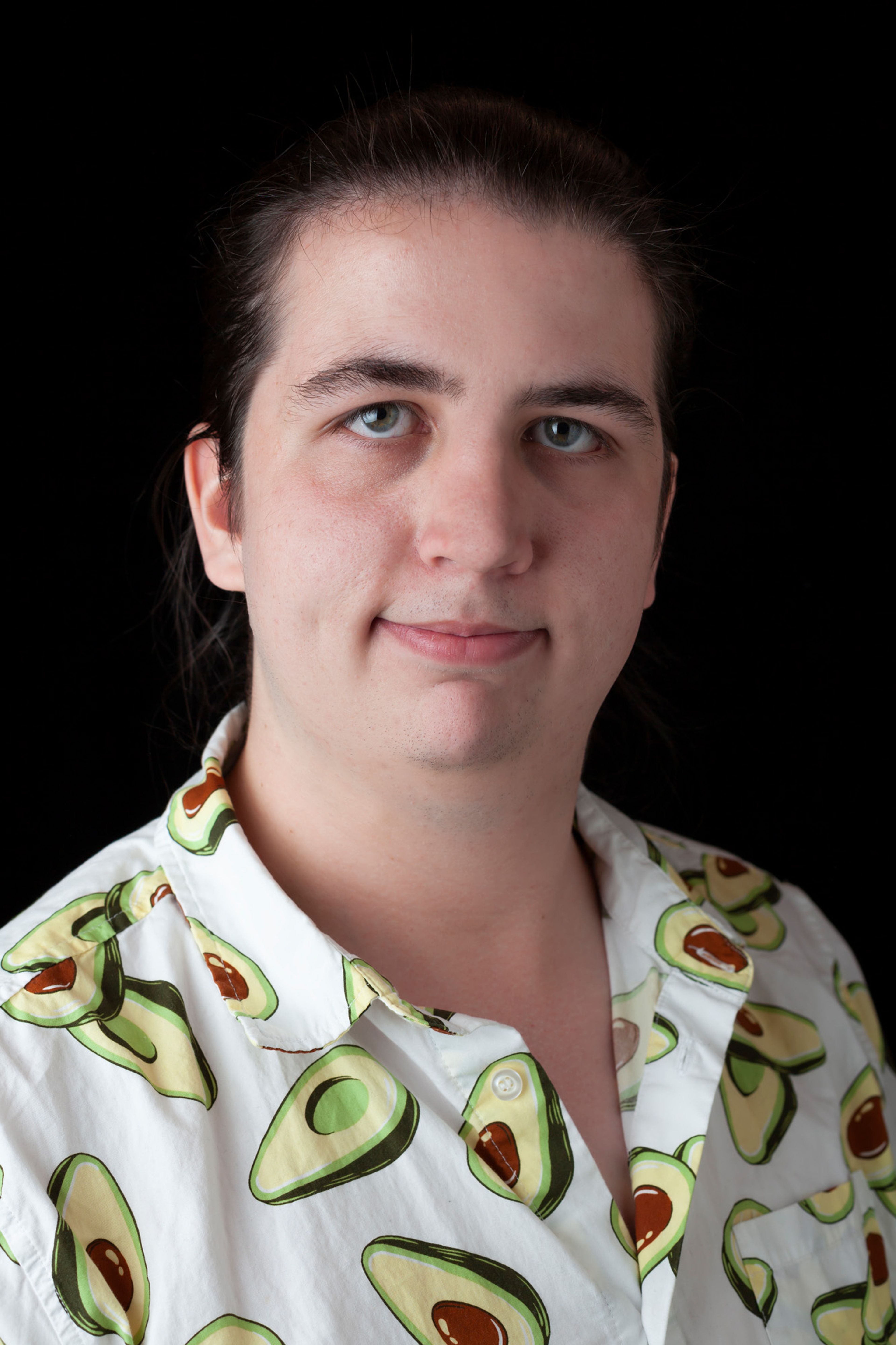 A person with long, dark hair tied back and a calm expression, wearing a white shirt adorned with an avocado pattern