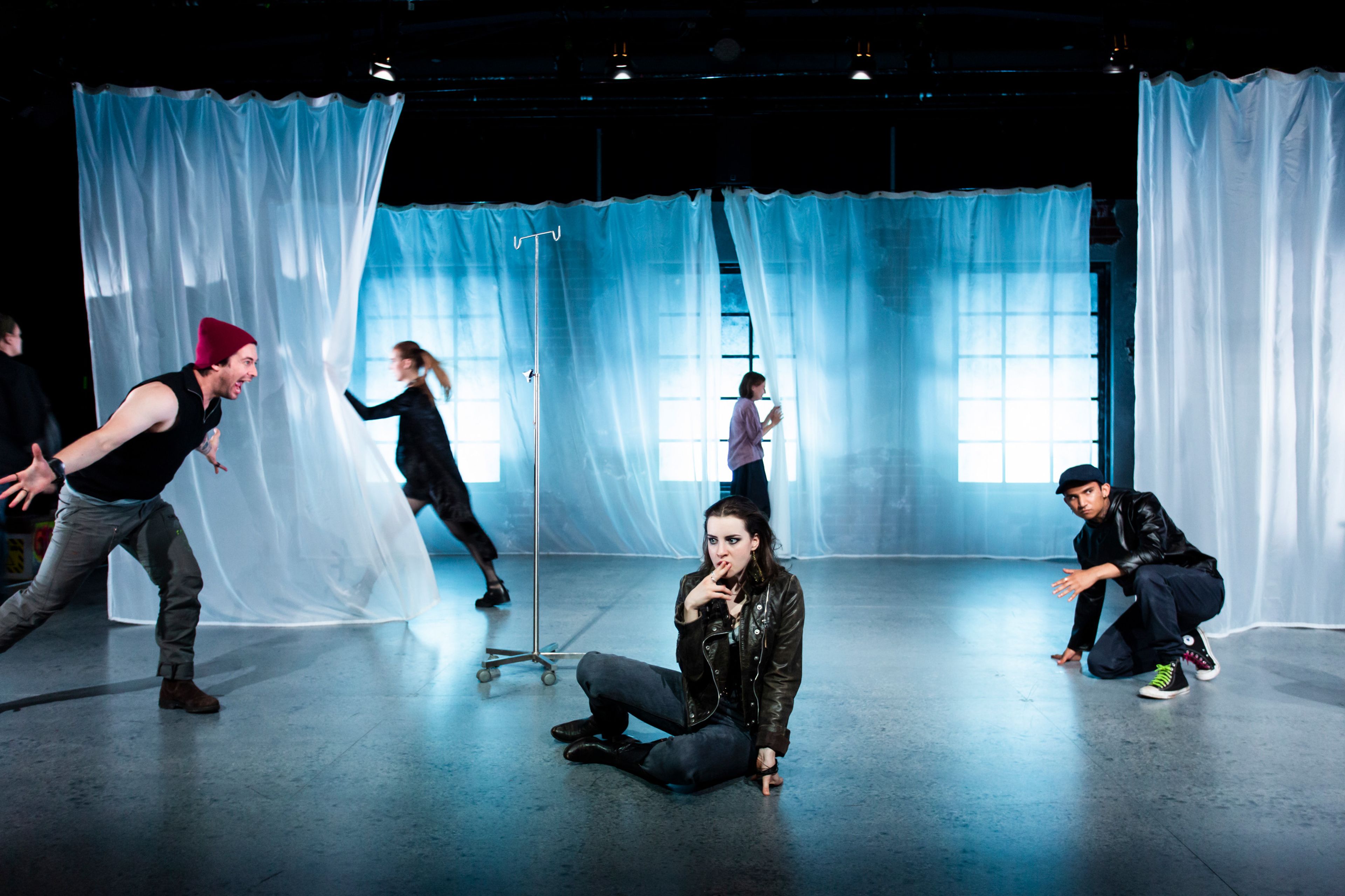 Performers in dynamic poses on a minimalist, blue-lit stage. A woman sits pensively in the foreground, while others move dramatically around translucent curtains and backlit windows.