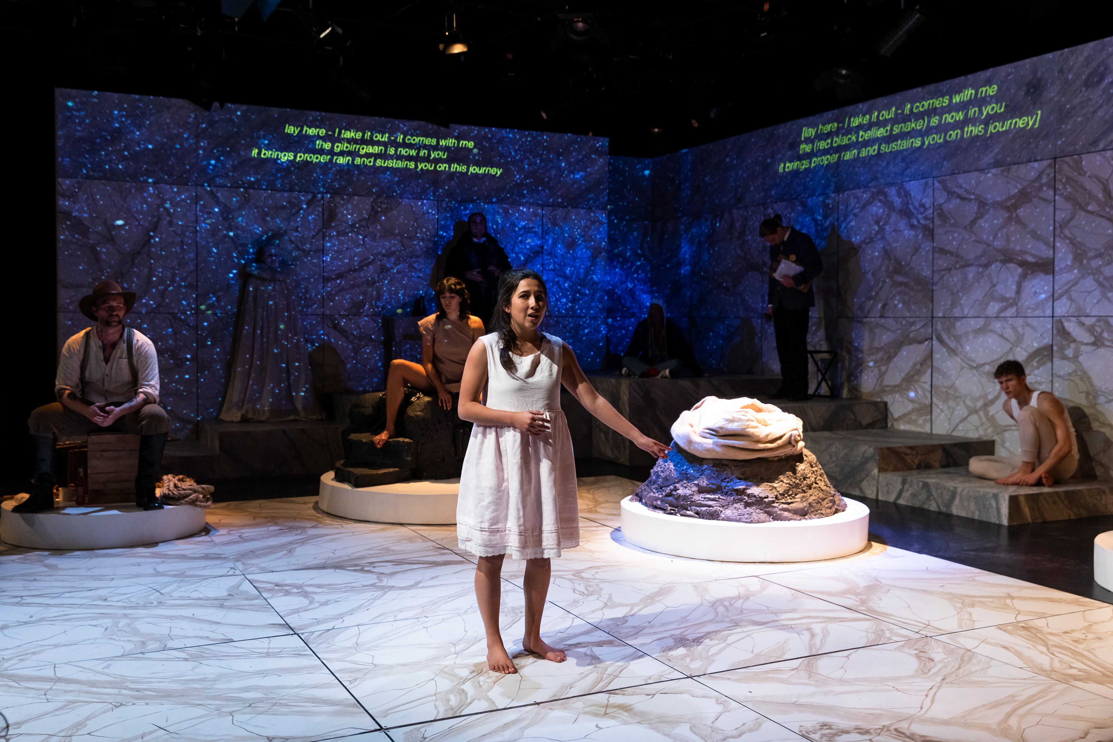 A barefoot performer in a white dress stands on a marble-patterned stage, pointing towards a rocky pedestal with fabric. Other actors, in varied costumes, are seated or posed in the dimly lit background against a starry projection, with subtitles displayed above