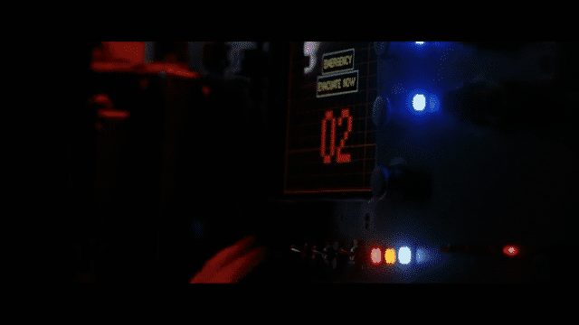 A dimly lit control panel with a glowing digital display reading "02" under the heading "EMERGENCY - DISENGAGE NOW," surrounded by blue and red indicator lights. A hand reaches toward the switches, adding a sense of urgency