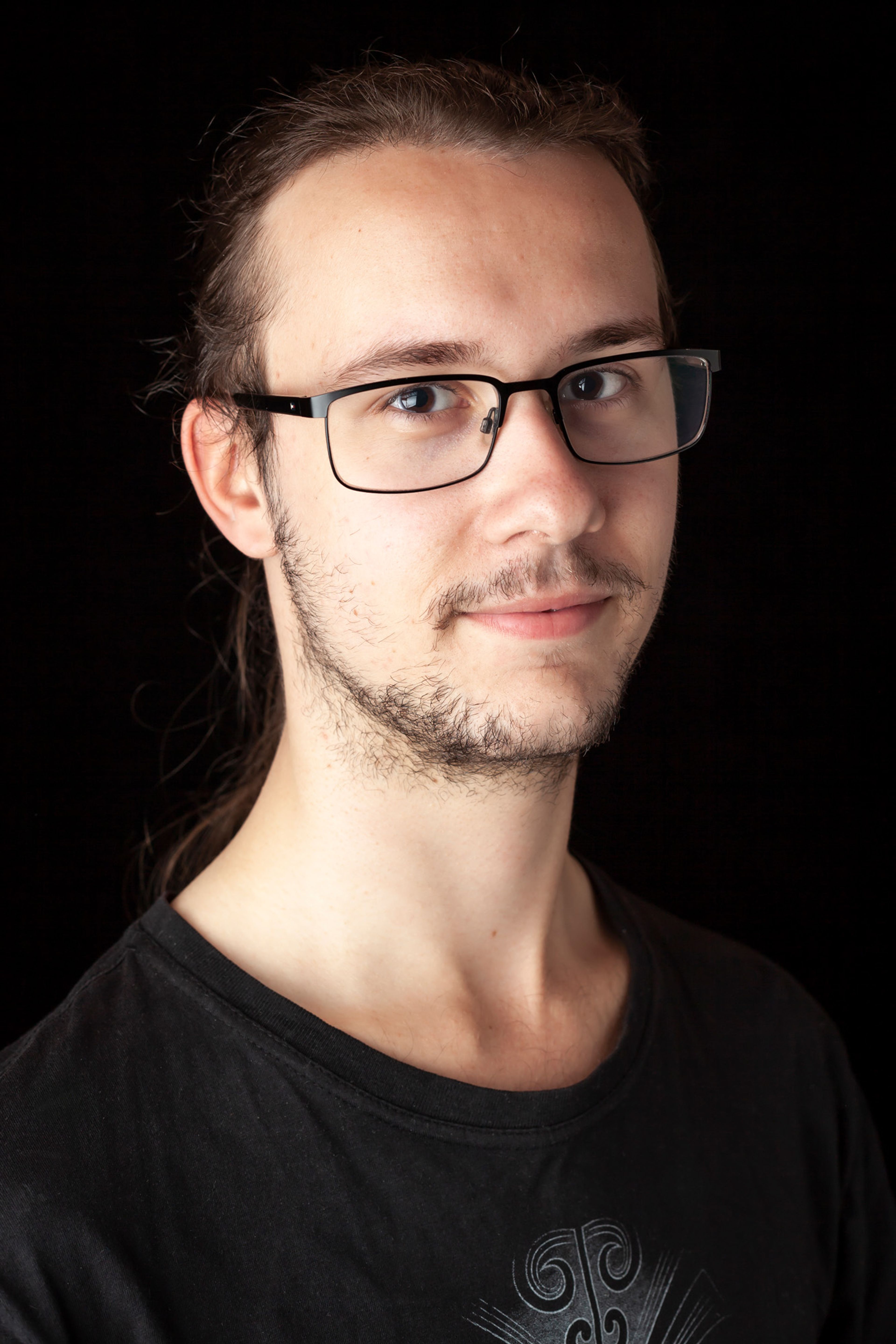 A portrait of a man with glasses, long tied-back hair, and light facial hair, wearing a black t-shirt